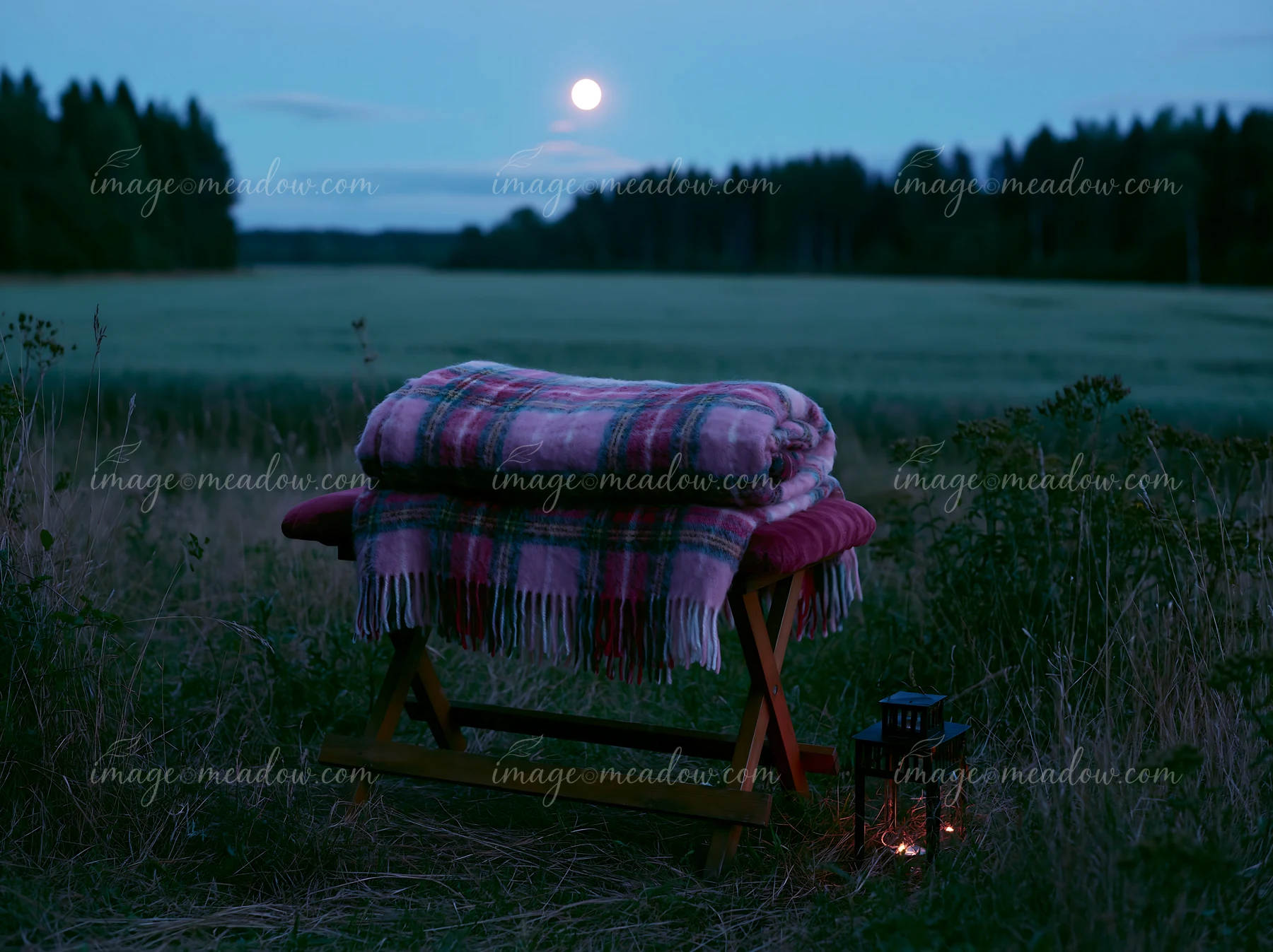 A blanket rests on a wooden bench in a meadow beneath a full moon.