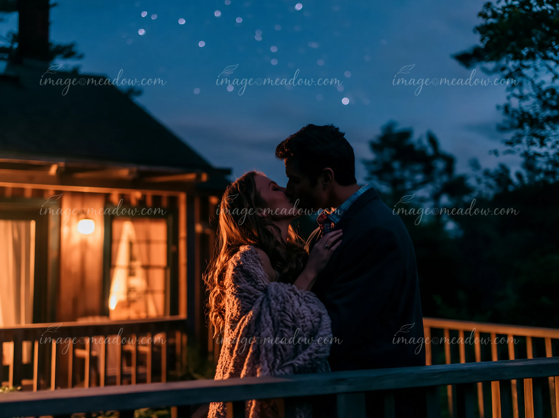 A couple kisses in front of a cabin surrounded by trees at night.