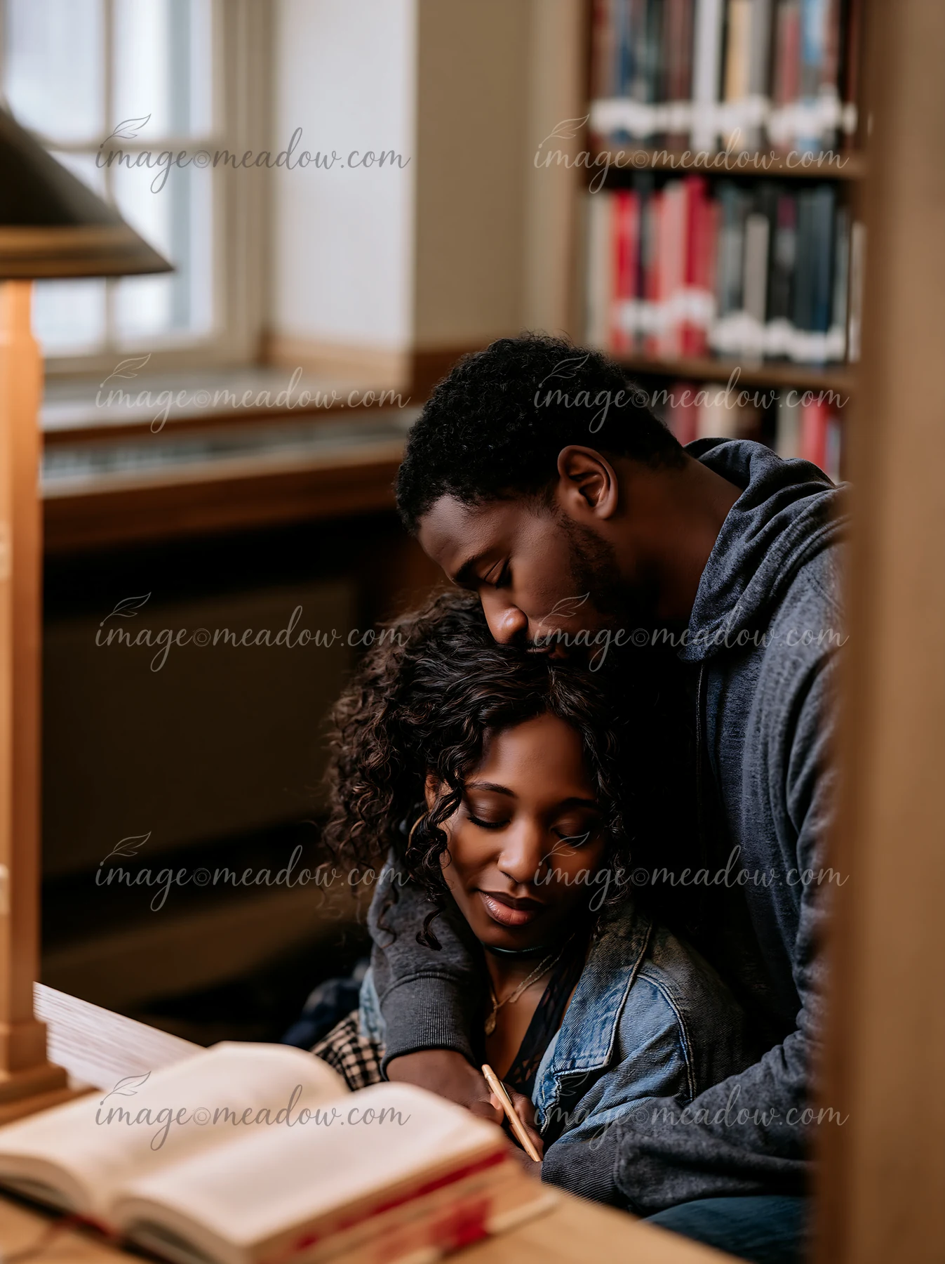 A couple shares a kiss on the forehead while sitting close together in a quiet library.