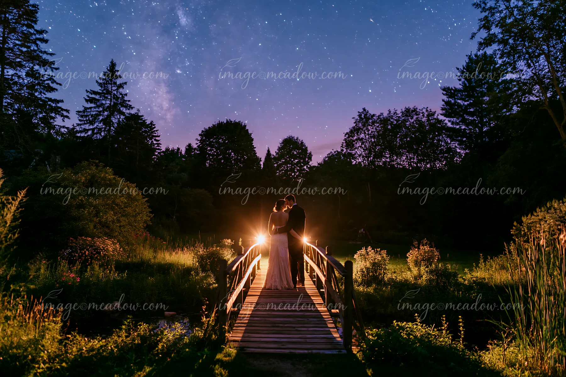A couple stands on a wooden bridge, sharing a moment under a starry night sky in a rural backyard.