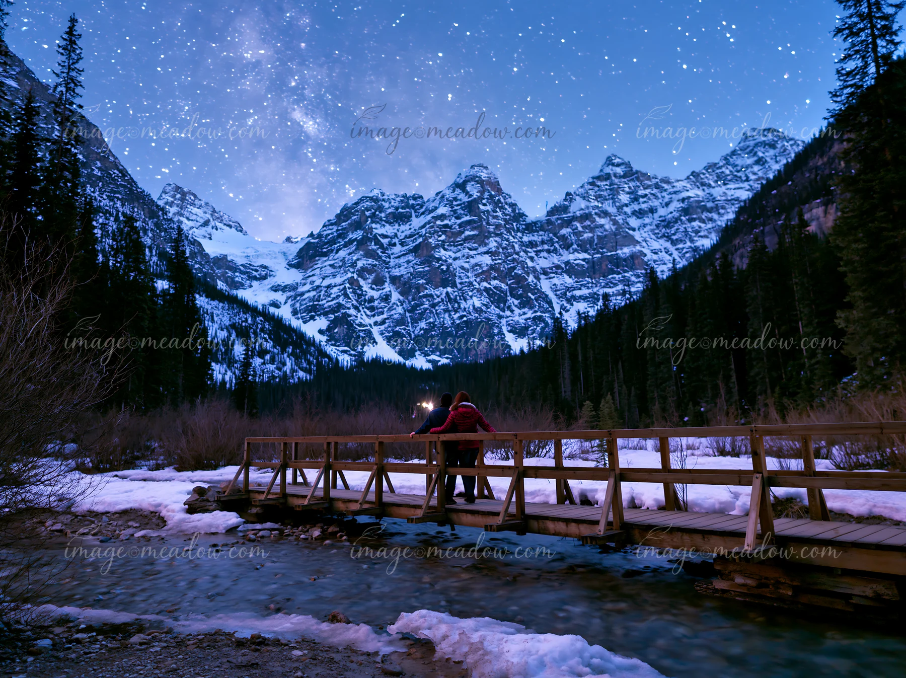 A couple under stars in snowy mountains stands on a wooden bridge over a stream.