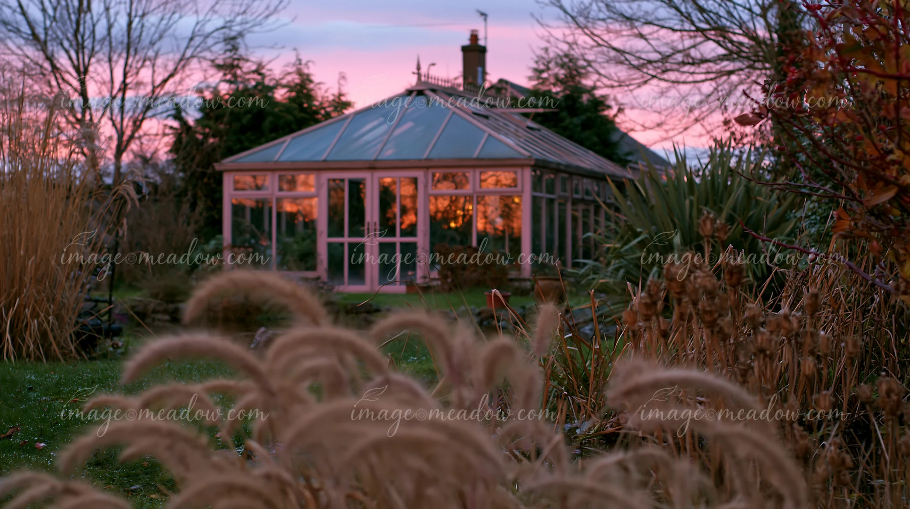 A glass garden conservatory stands surrounded by plants and grass under a colorful evening sky.