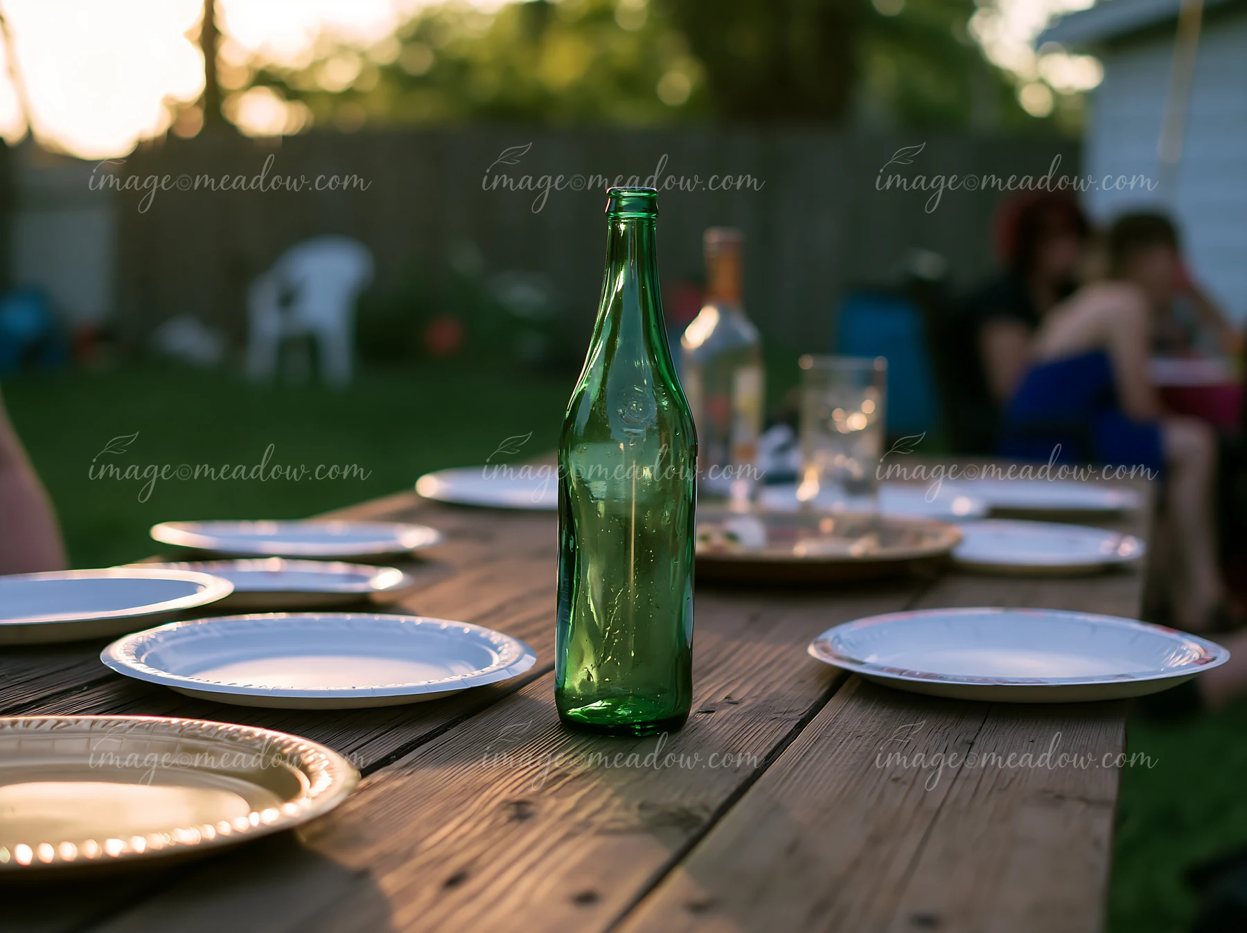 A green empty bottle stands on a wooden table surrounded by plates during a backyard gathering.