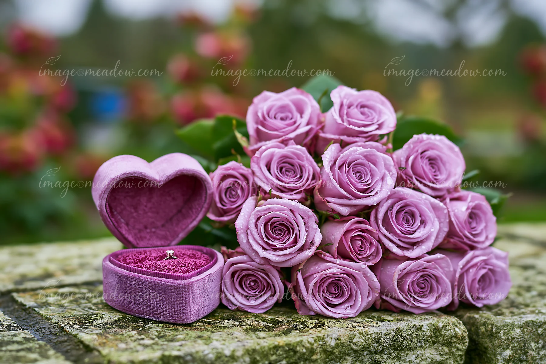 A heart-shaped ring box sits beside a bouquet of pink roses on a stone surface.