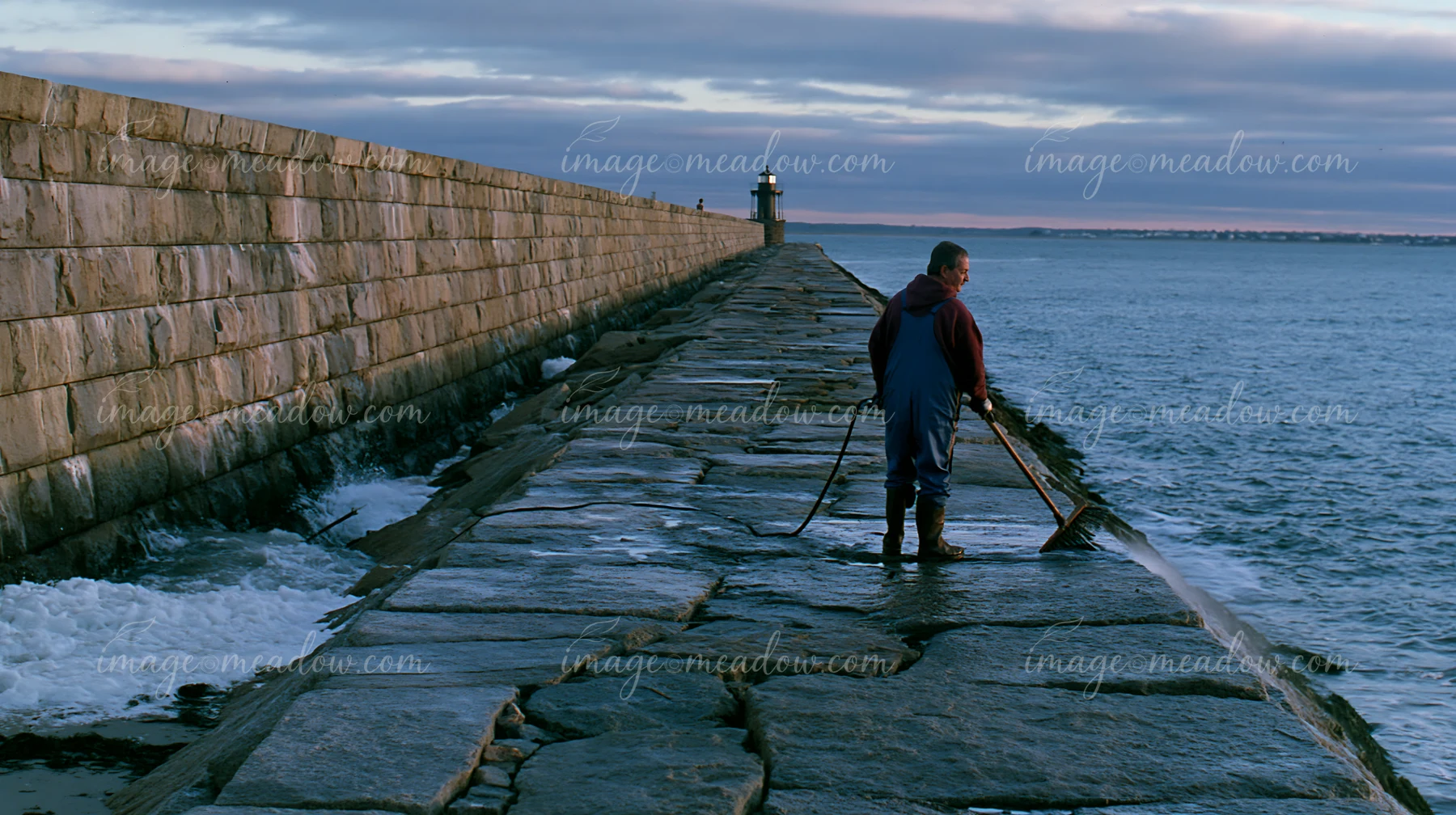 A lighthouse maintenance worker at work stands on a stone pier, using tools to maintain the structure as waves crash nearby.