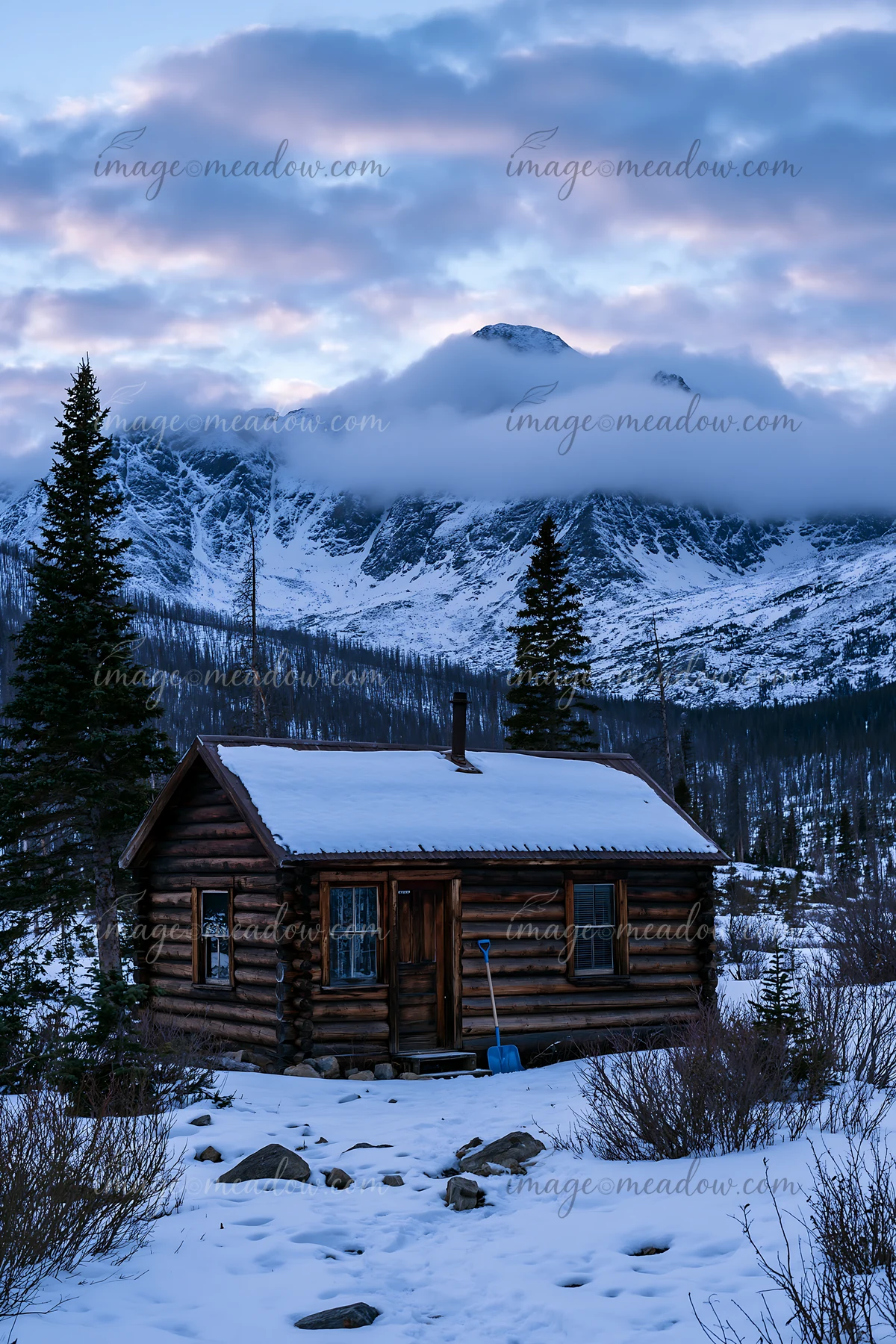 A log cabin stands in the snow with dramatic clouds around alpine peaks in the background.