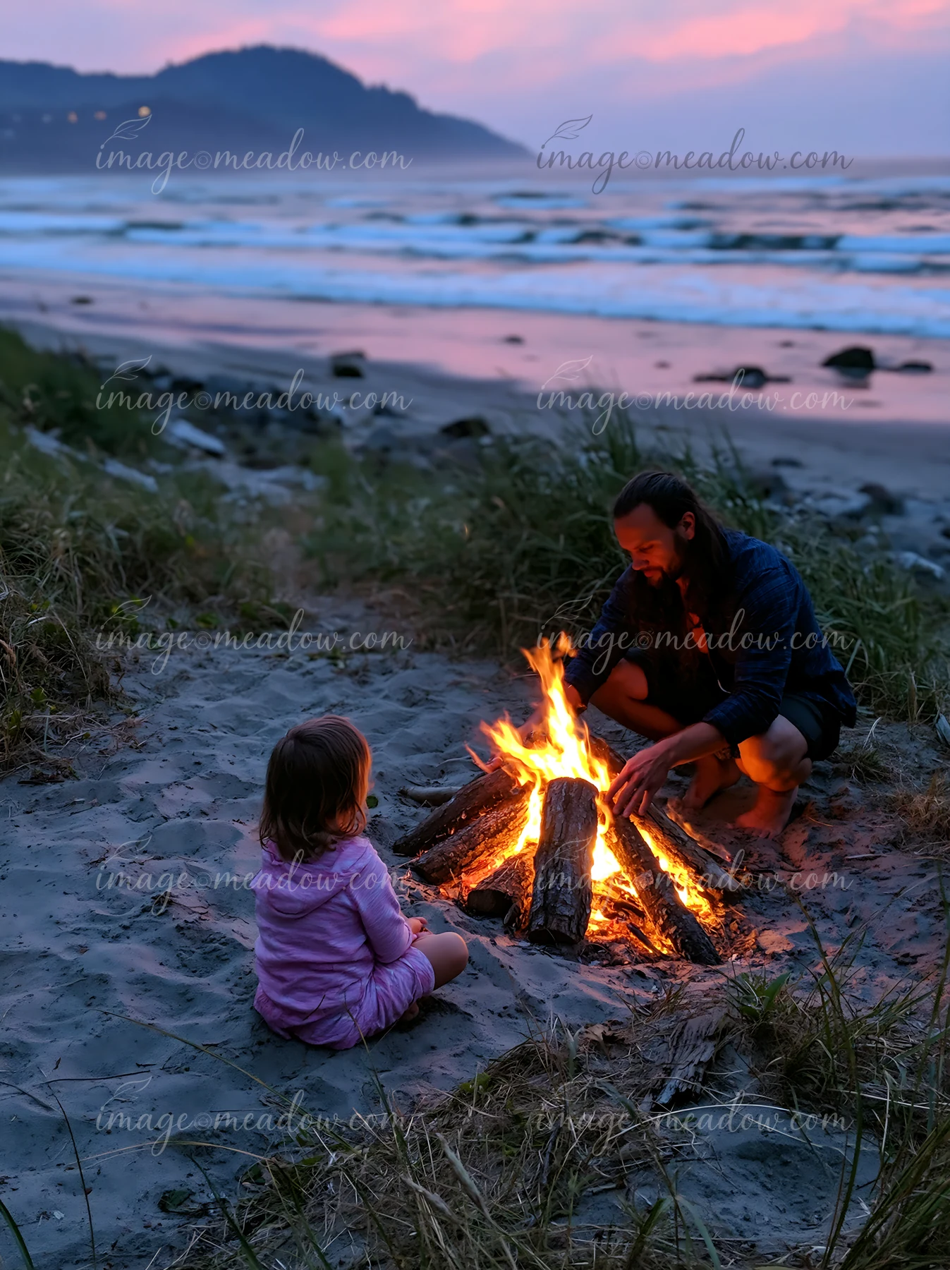 A man builds a fire while a young girl sits nearby on a sandy beach at twilight.