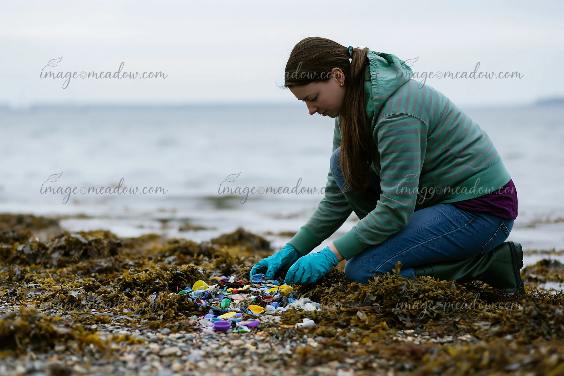 A person collects plastic waste from a rocky coastline covered in seaweed.