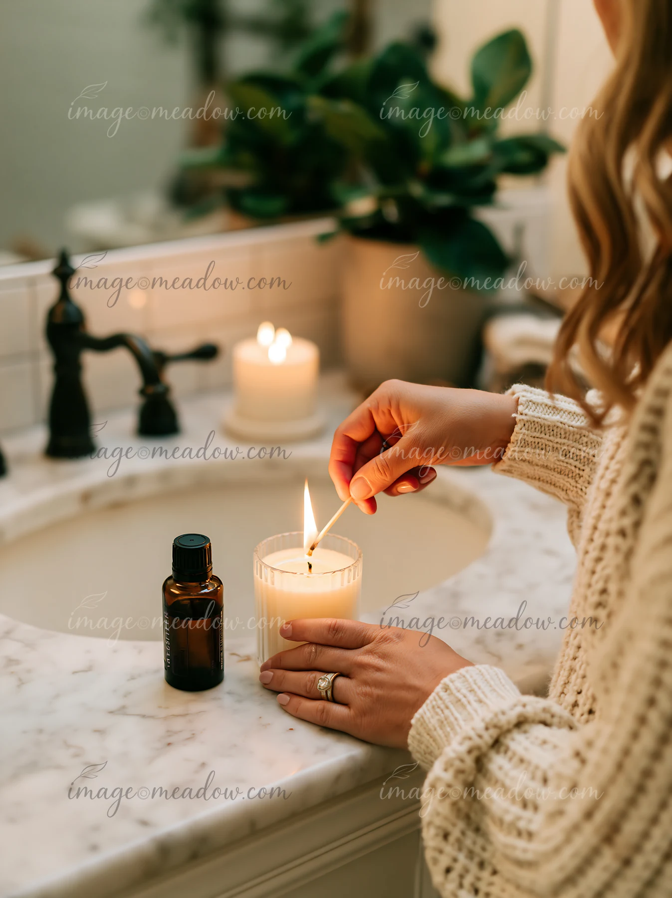 A person lights a candle next to an essential oil bottle on a marble bathroom counter.