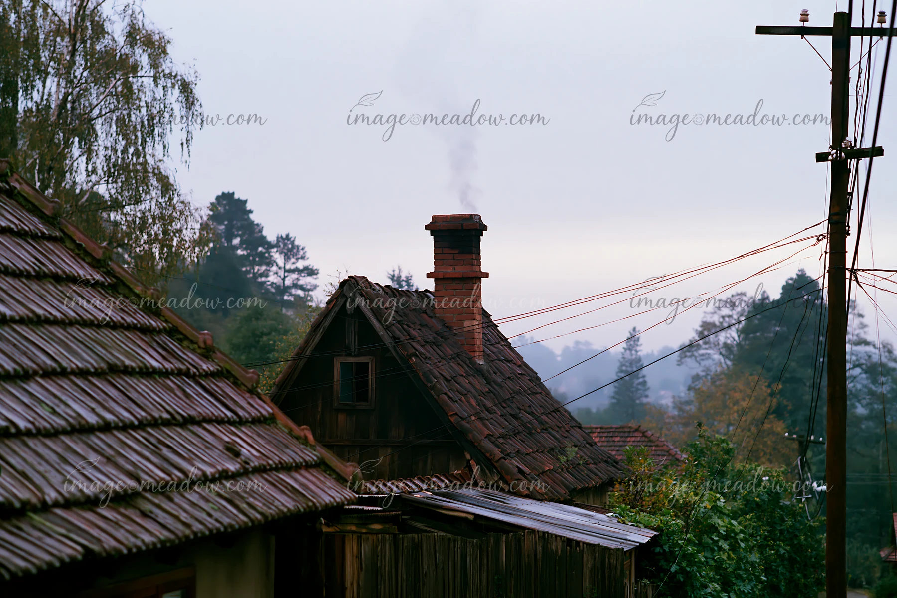A rural cottage after rain features a chimney with smoke rising, surrounded by trees and power lines.