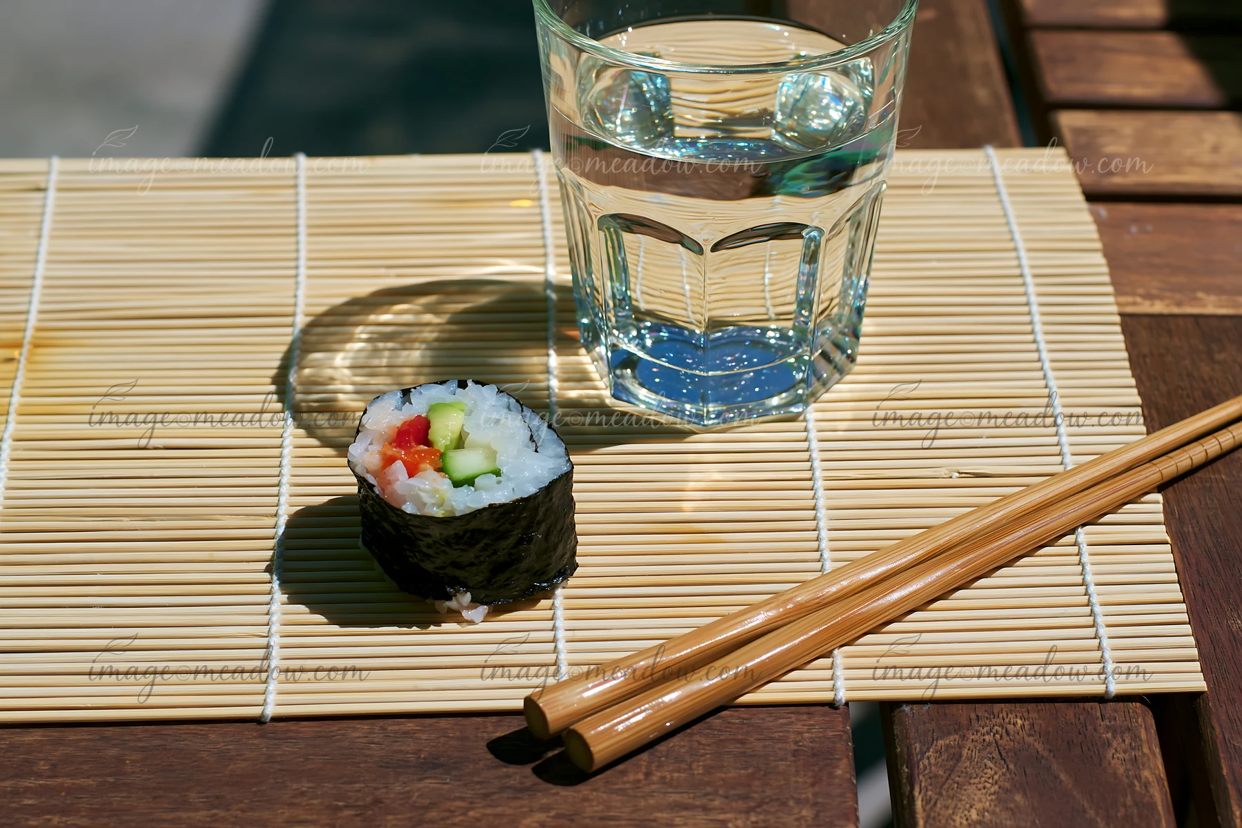A single sushi roll on a bamboo mat sits next to a glass of water and chopsticks.