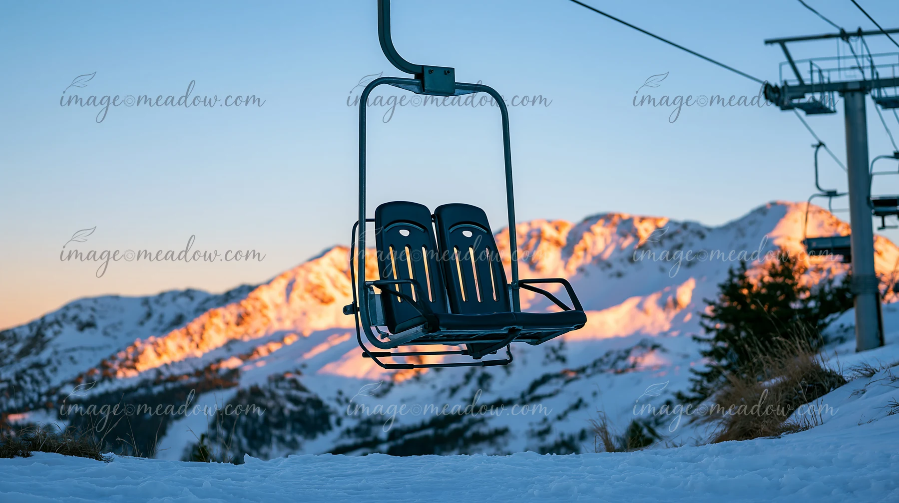 A ski lift stands empty against snowy peaks illuminated by golden sunlight at sunset.