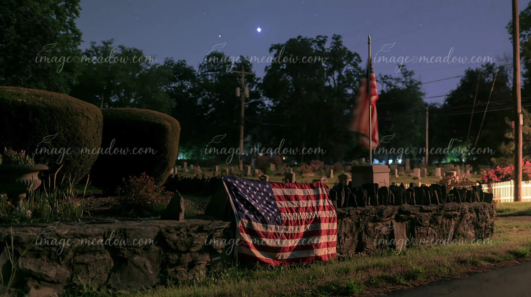 A solemn cemetery scene with American flag and stars shows a flag draped on a stone wall among gravestones under a night sky.