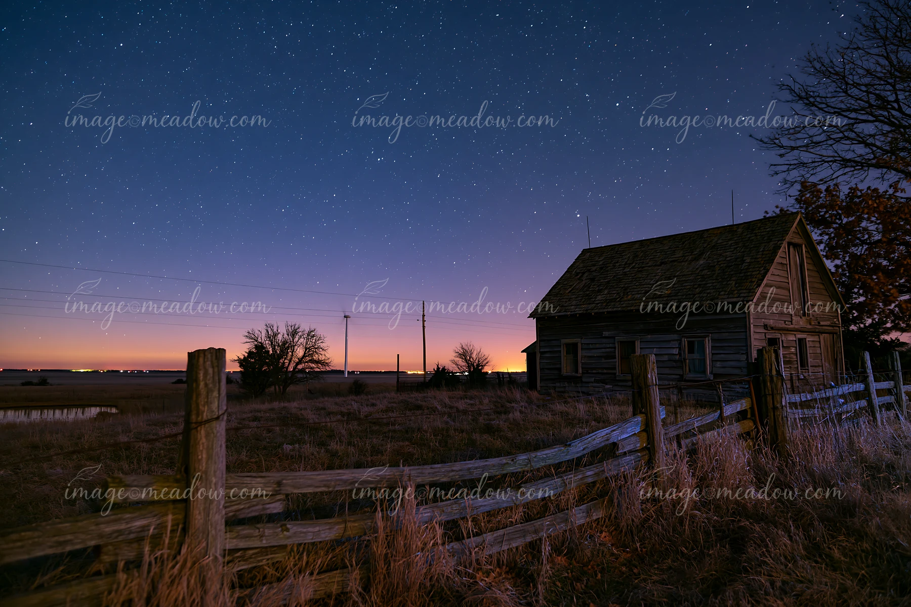 A starry night above a rural wind turbine and a rustic farmhouse shows a clear sky filled with stars.