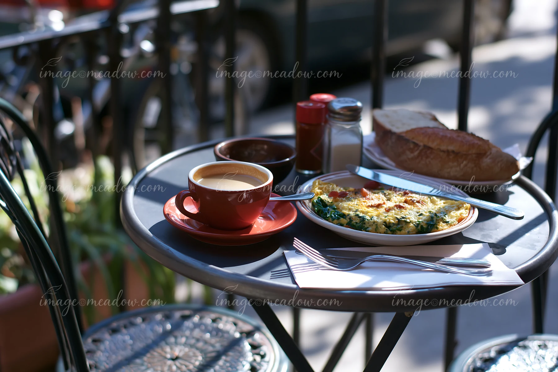 A street cafe table holds a cup of milk coffee alongside a plate of breakfast food.