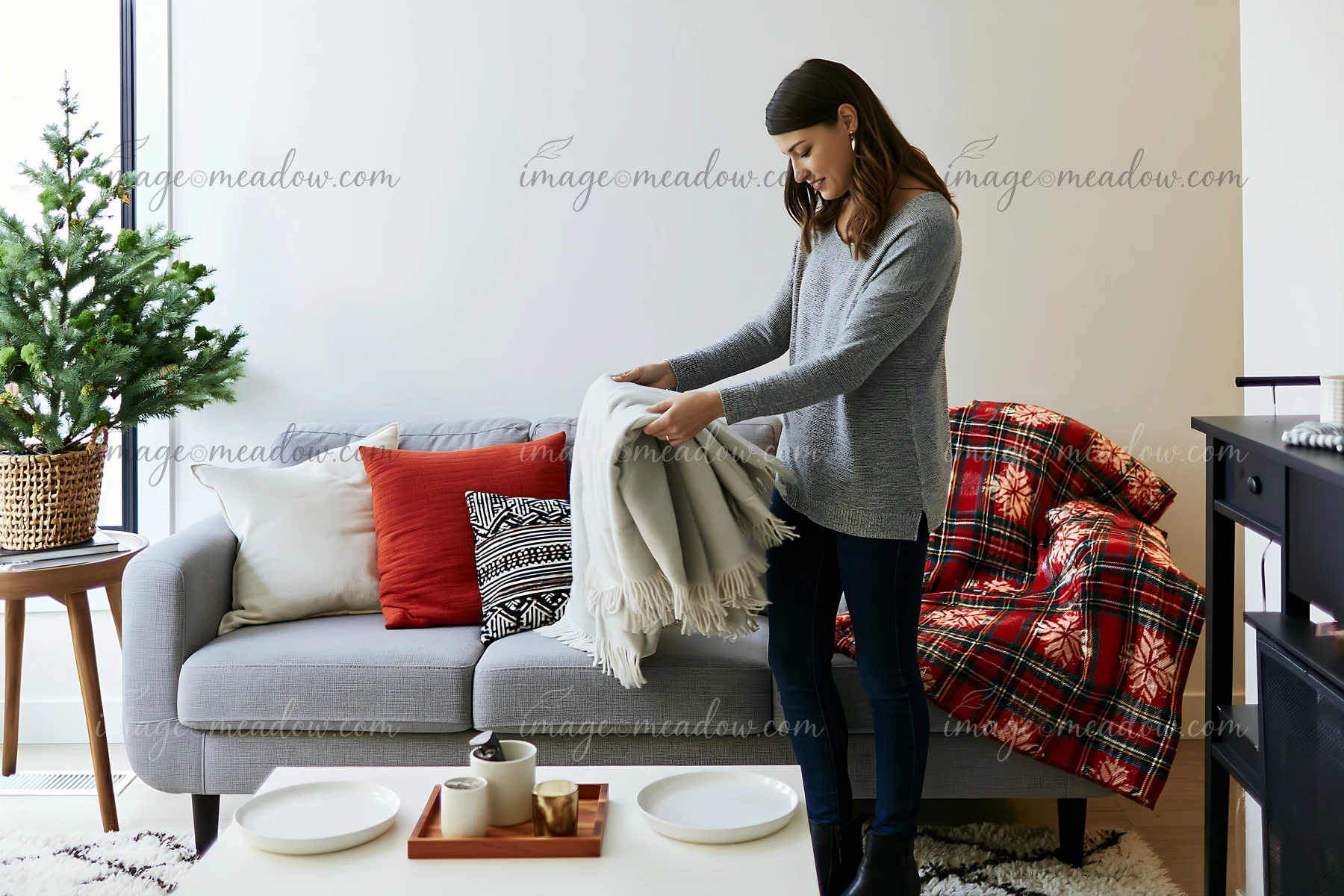 A woman arranges a blanket on a festive sofa with pillows in a modern living room.