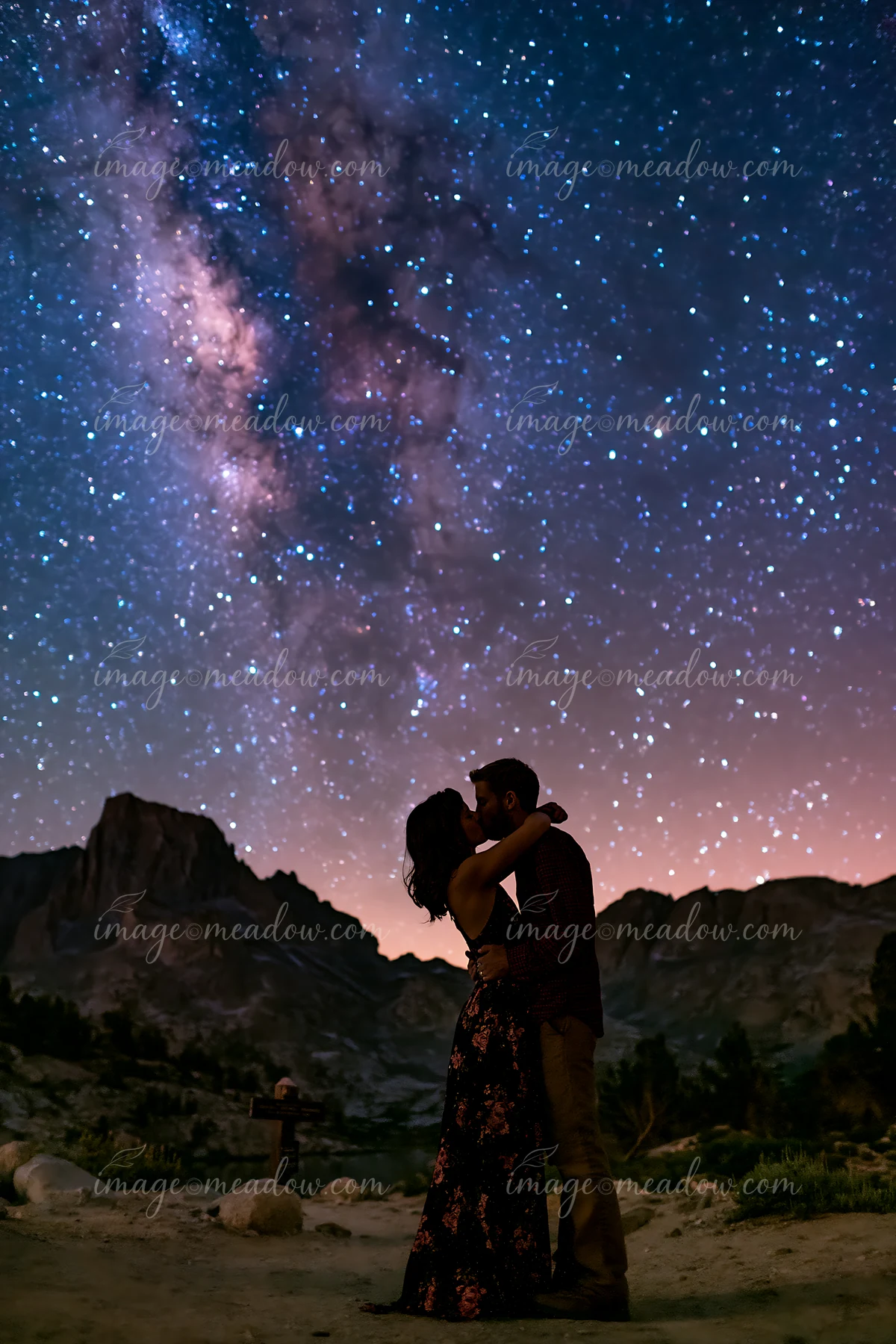 A young couple kissing beneath visible Milky Way while surrounded by mountain scenery at night.