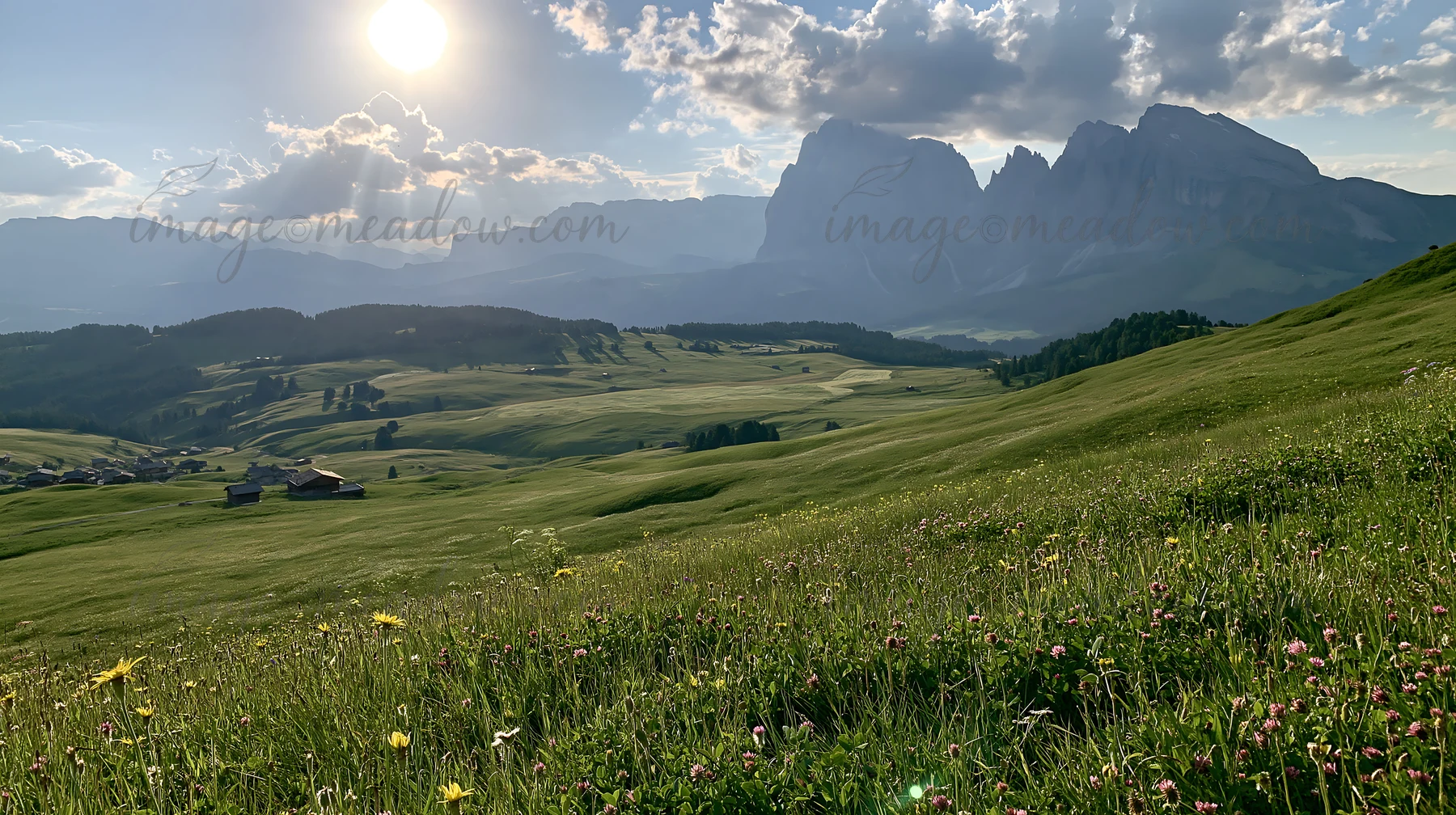 Sunbeams shine over the lush Alpe di Siusi meadow. Green grass covers the area, and distant mountains rise sharply against a cloudy sky. This scene captures the