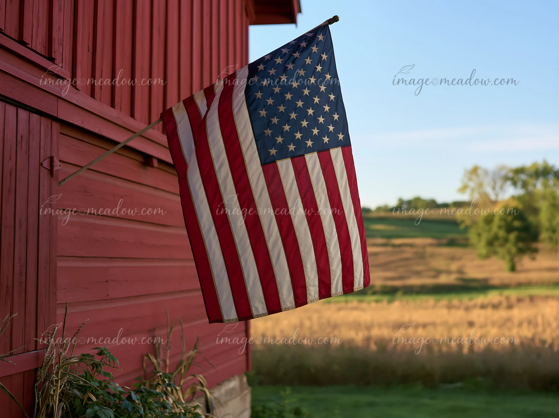An American flag waving in gentle breeze hangs from a rustic barn in a rural setting.