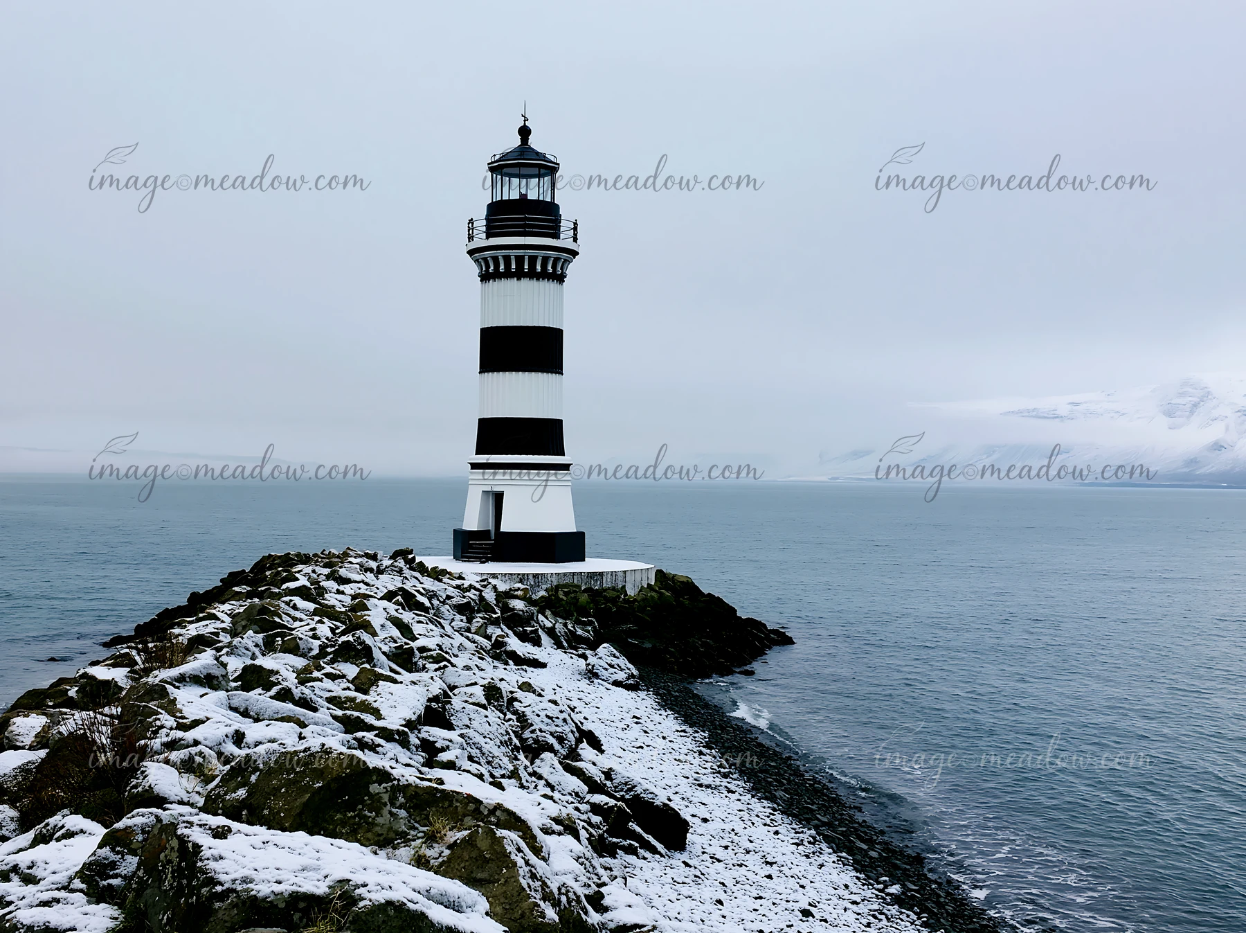 Black and white striped lighthouse stands on a snowy rocky coast beside calm water and misty snow-covered mountains.