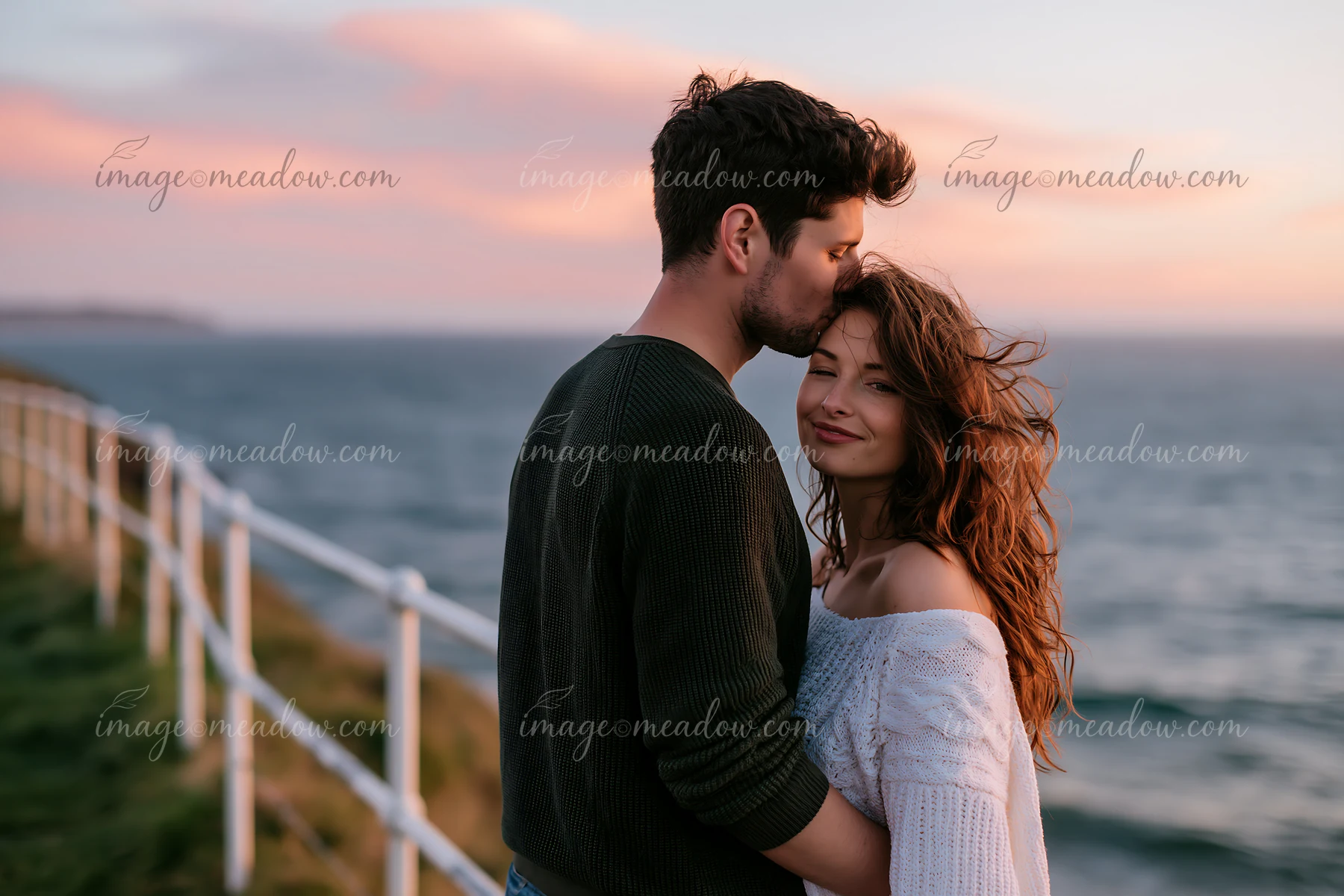 Couple sharing a forehead kiss beside the sea at sunset with a coastal railing behind them.