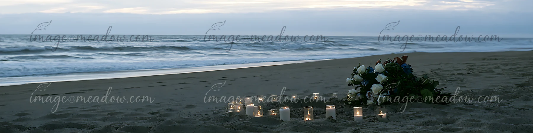 Flowers and lit candles form a memorial on a sandy beach beside the ocean.