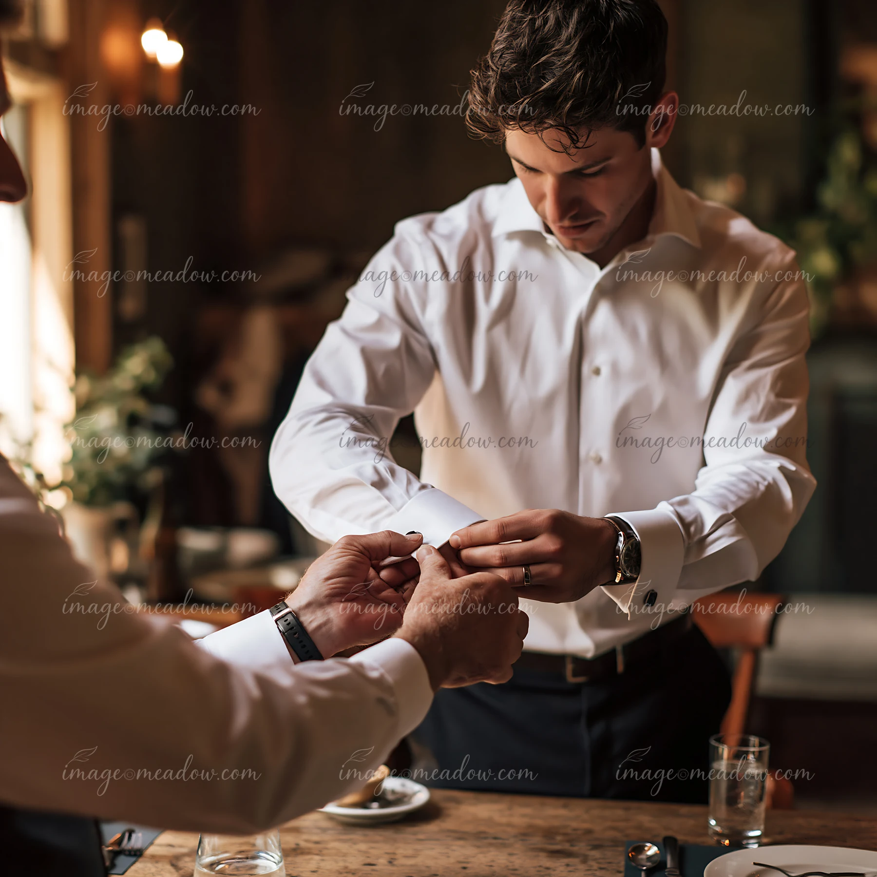 Groom adjusts his cufflinks with help from another person in a warmly lit room.