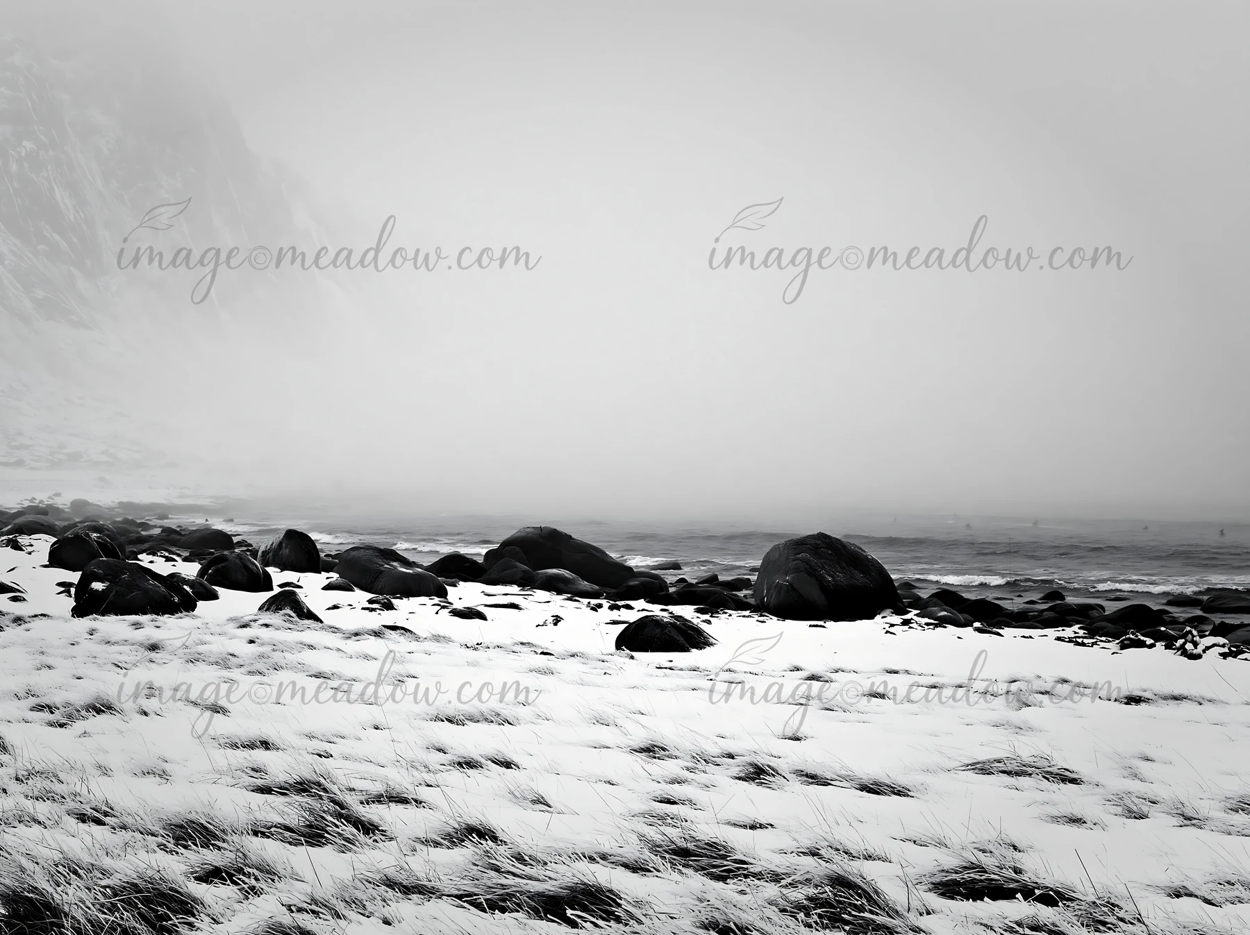 Lofoten coastal meadow covered in snow with rocks scattered along the shoreline. Foggy weather adds a gray atmosphere to the landscape during winter.