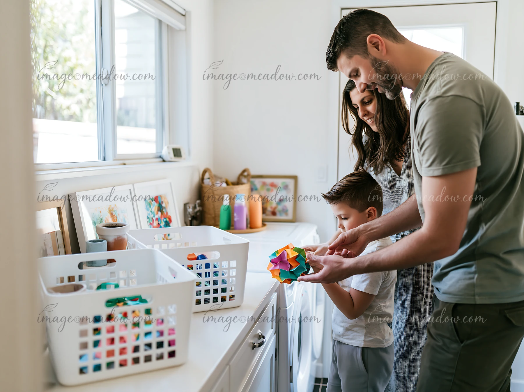 Parents and their young son examine colorful artwork together in a bright home laundry room.