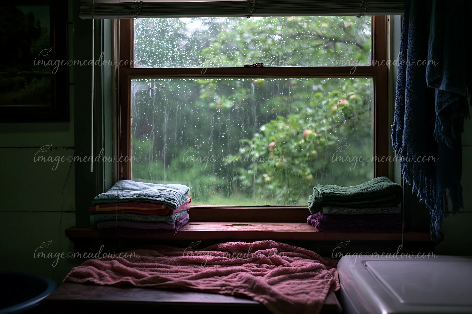 Raindrops cover a wooden window in a rural home, with fresh towels stacked on the sill.