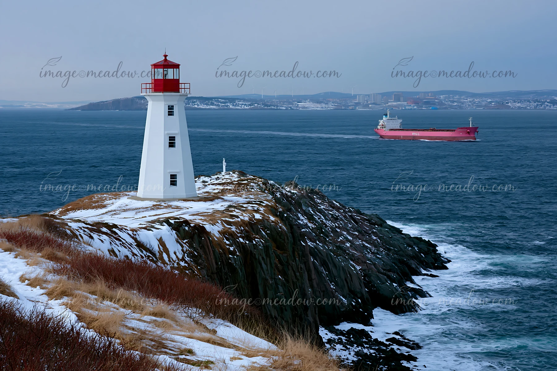 Red-and-white lighthouse on a snowy rocky coast with a cargo ship sailing in the distance.