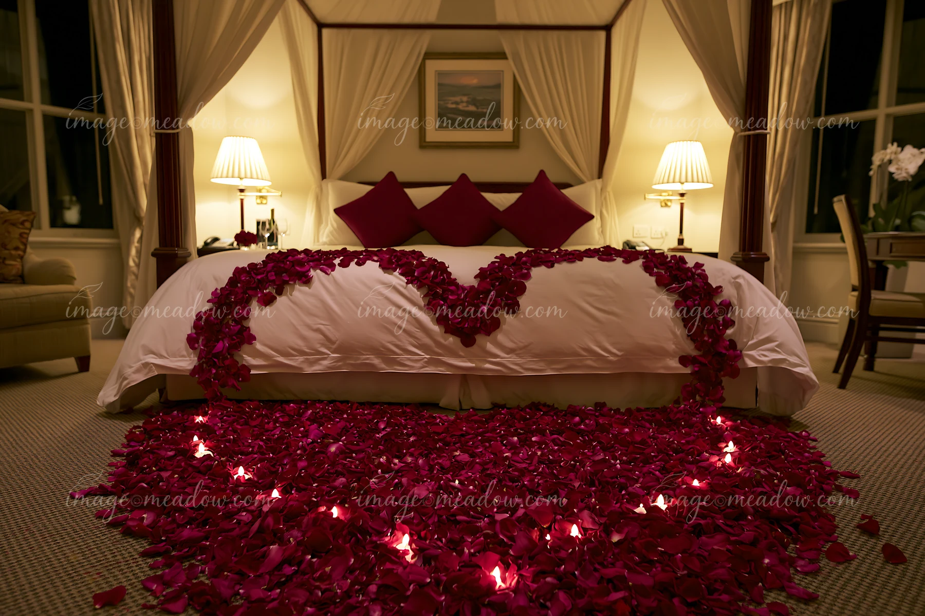 Romantic bedroom with red rose petals forming hearts across a bed and floor, illuminated by candles and bedside lamps.