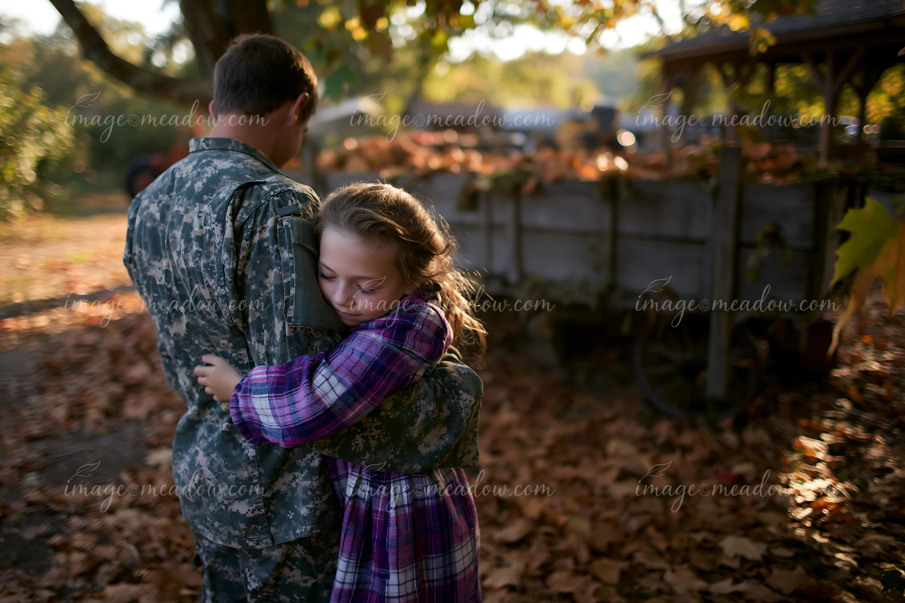 Soldier in camouflage embraces a young child among fallen leaves and rustic outdoor structures.