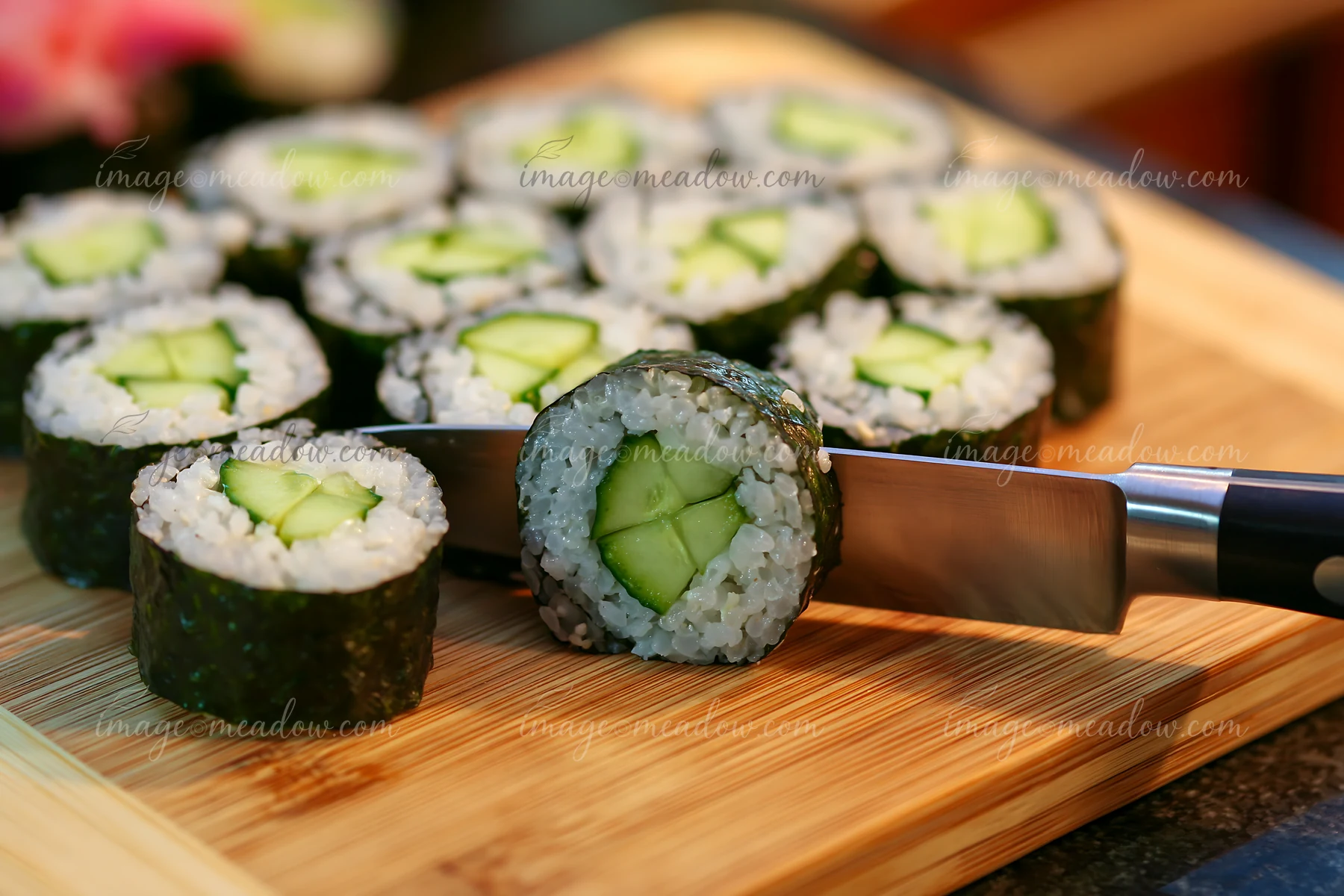 Sushi rolls being cut with knife on a wooden cutting board in a kitchen.