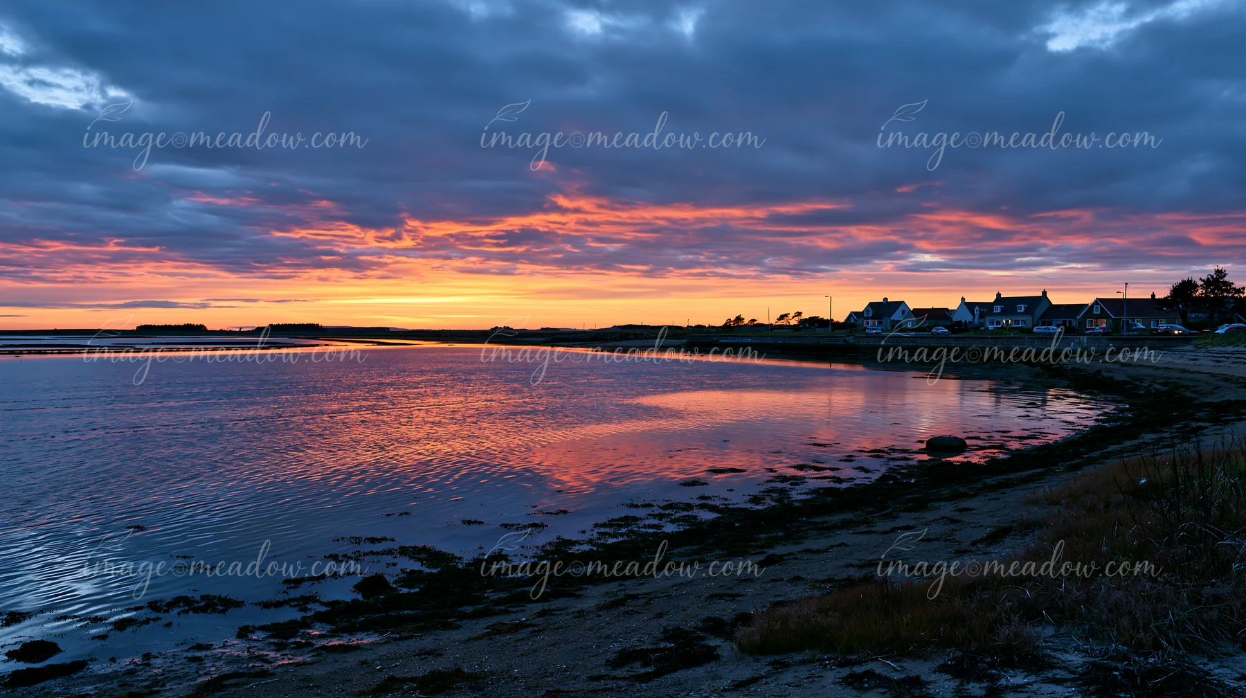 The view shows a coastline with mussel farms at sunset, with reflections in the water and houses along the shoreline.