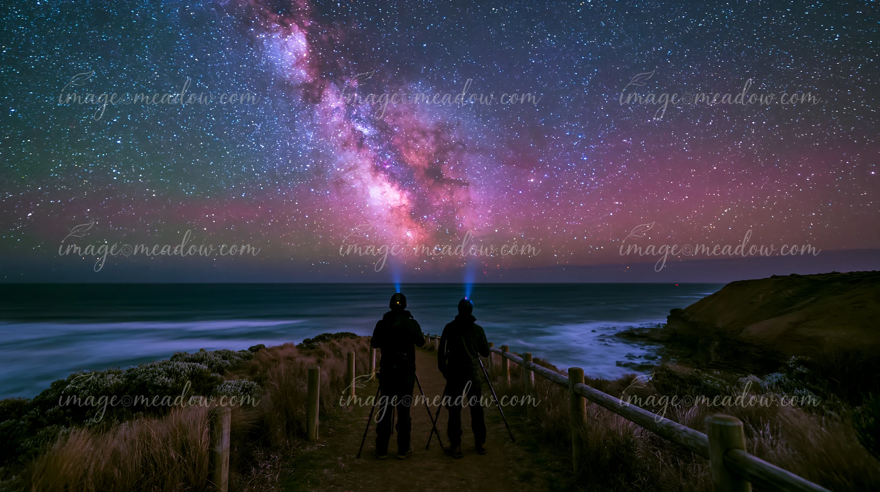 Two people stand on a coastal path, using tripods to capture the Milky Way.