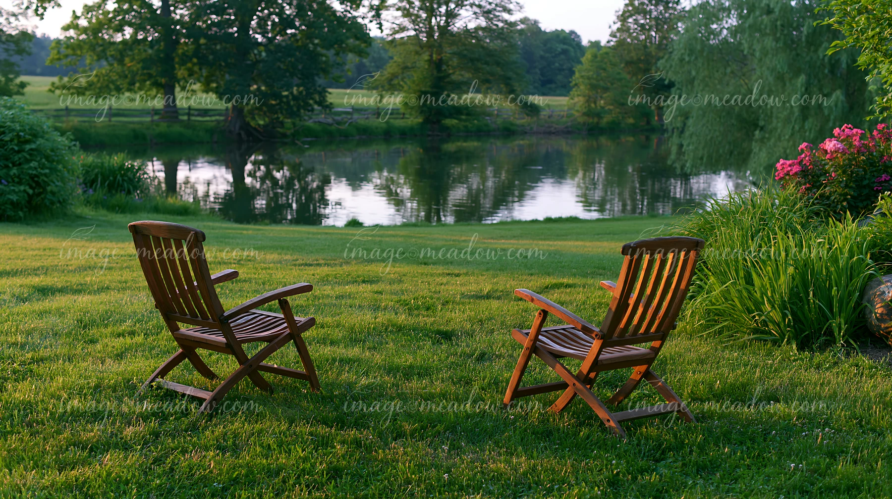 Two wooden chairs sit in a freshly mowed meadow facing a calm river surrounded by trees.