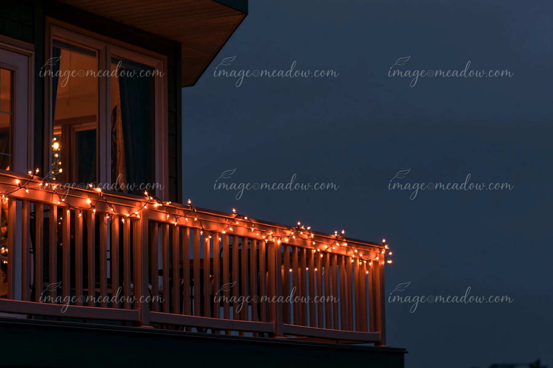 Warm Christmas lights glow along a residential balcony railing at dusk beneath a dark blue sky.