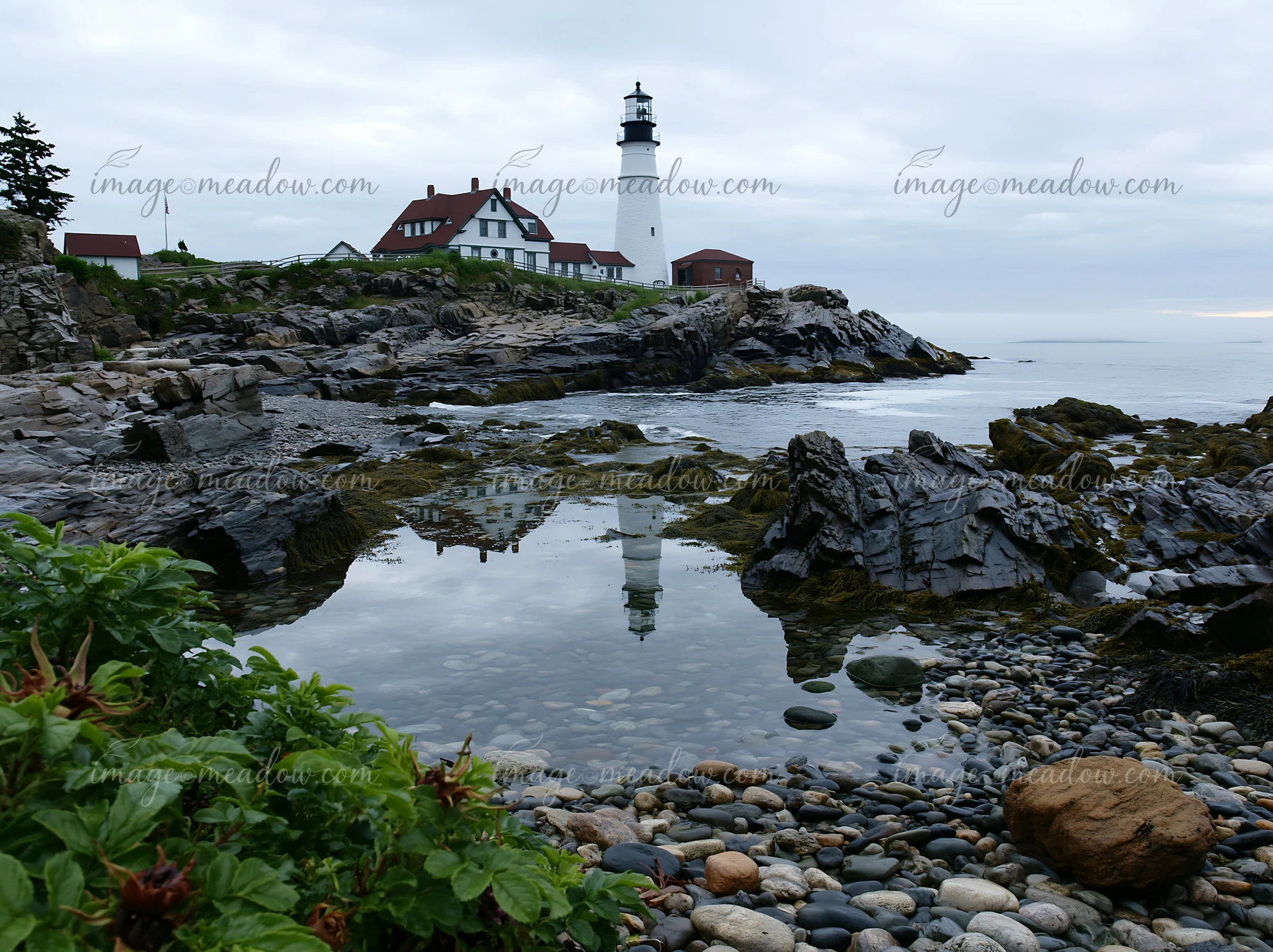 White lighthouse overlooking a rocky tide pool and pebble beach beneath an overcast coastal sky.