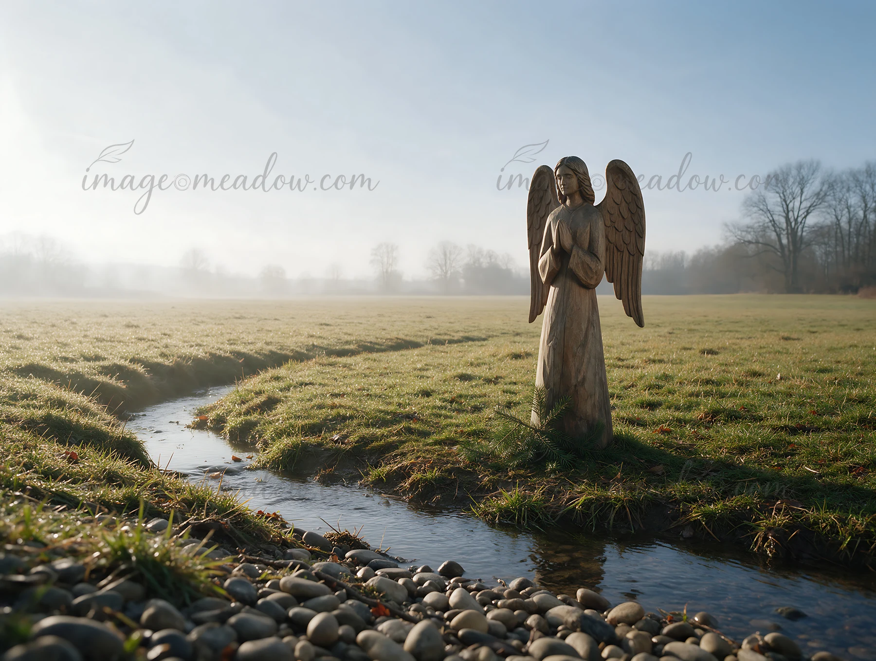 A wooden angel figure is positioned in a grassy meadow. A small stream flows nearby while fog covers the area, creating a quiet morning scene. The angel remains