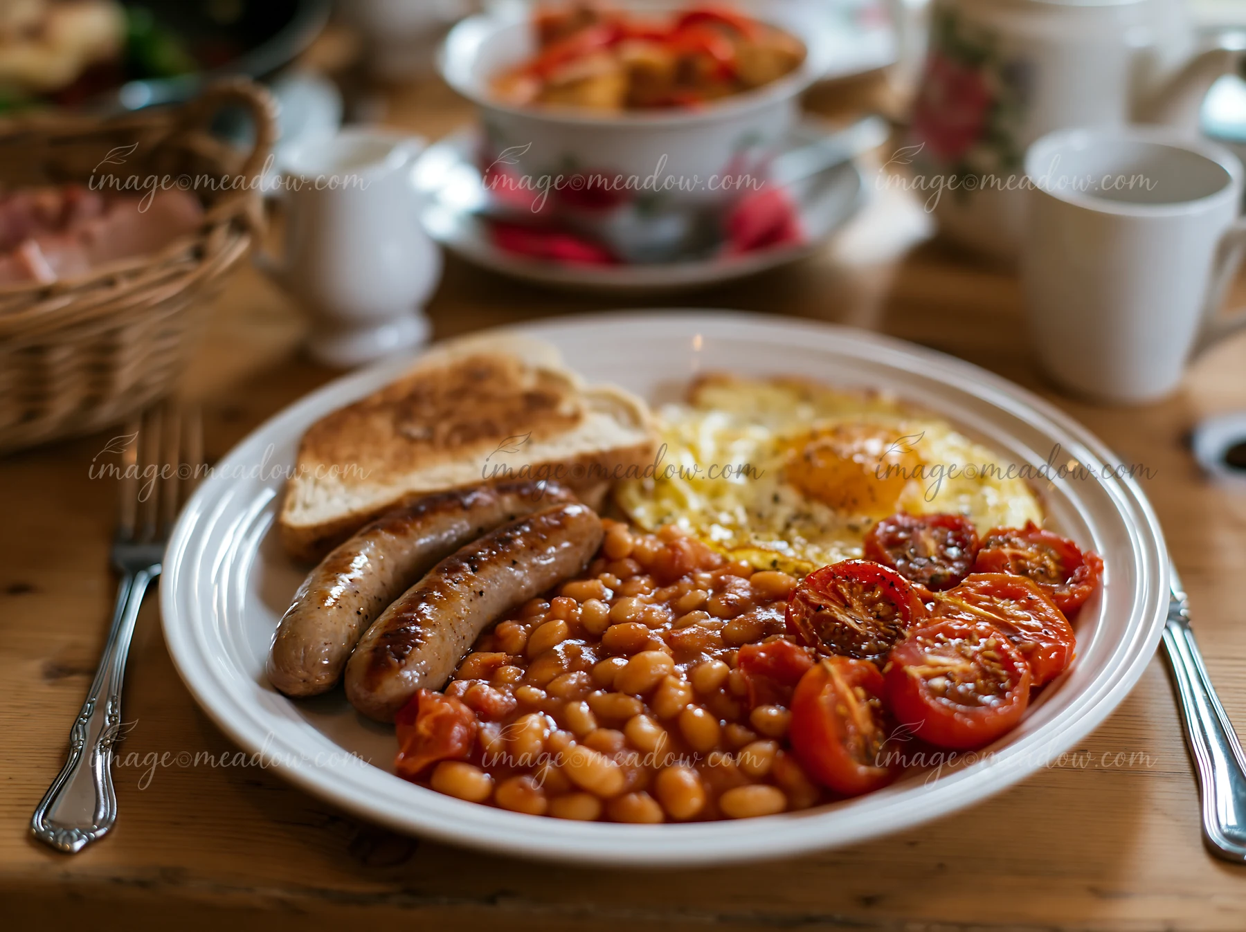 A breakfast plate with sausage, eggs, baked beans, and grilled tomatoes sits on a wooden table.