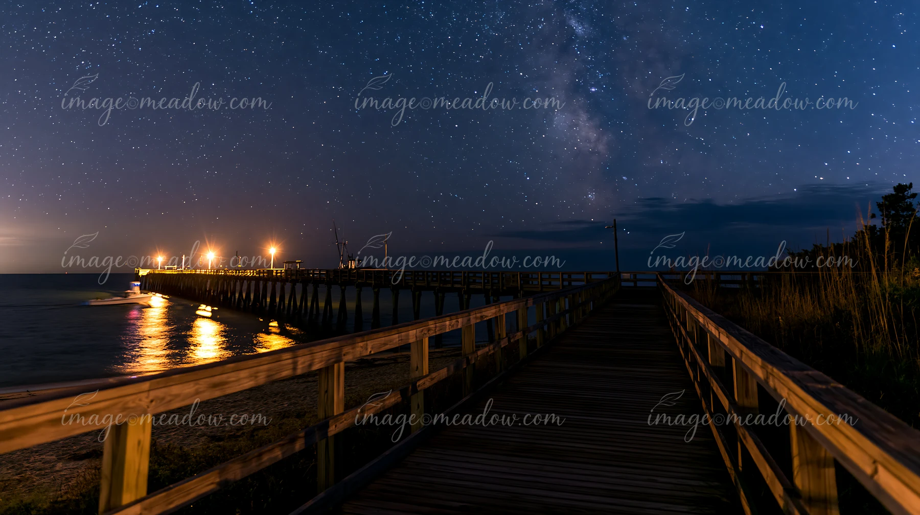 A bright starry night over a coastal boardwalk leads to a lit pier and ocean waves.