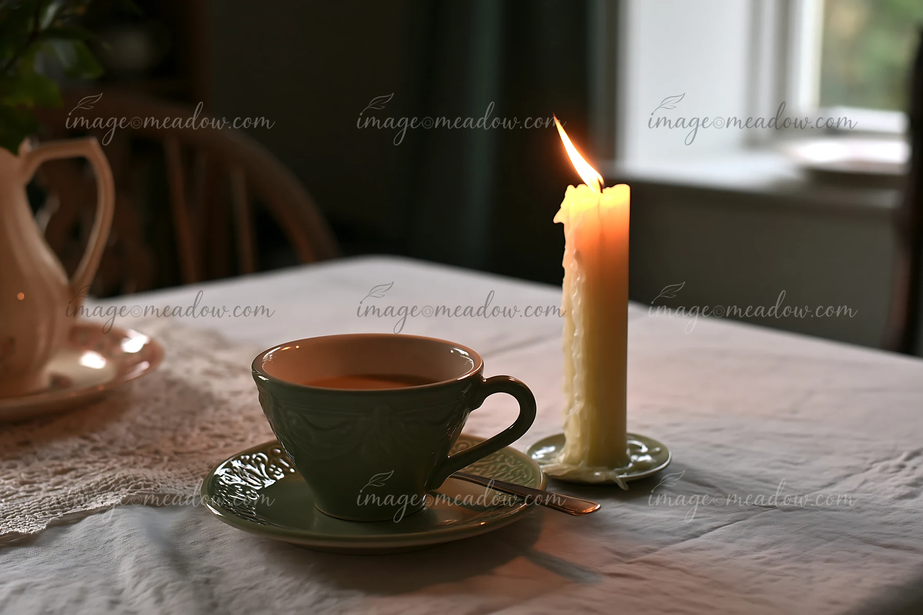 A candle with copy space on right stands next to a cup of tea on a table in a softly lit room.