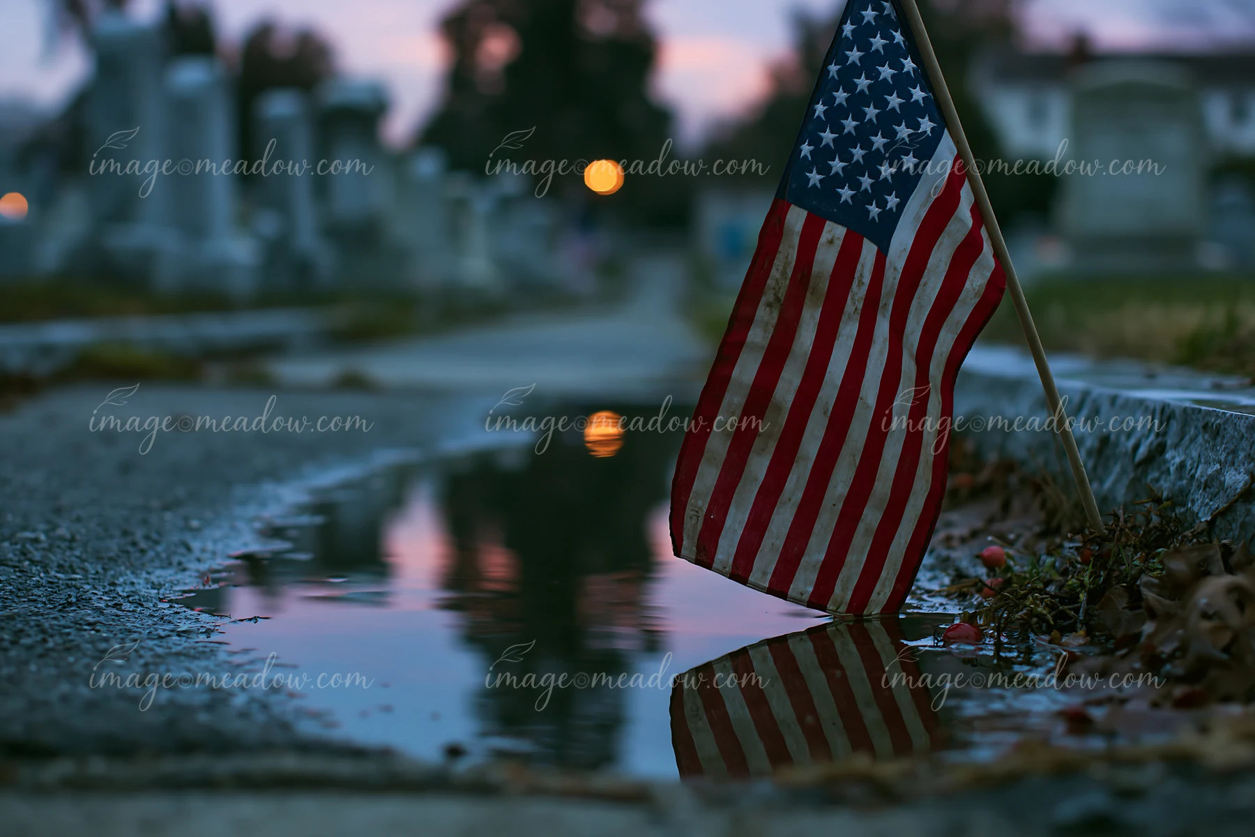 A close-up view of a small American flag at a veterans cemetery, reflecting in a puddle on a pathway as dusk settles over the gravestones in the background.