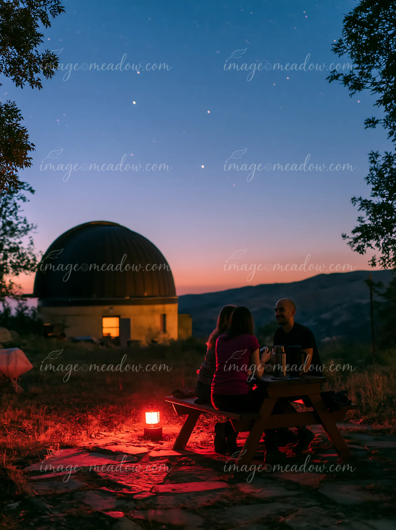 A couple at a remote observatory at night sits at a table under the stars, sharing drinks.