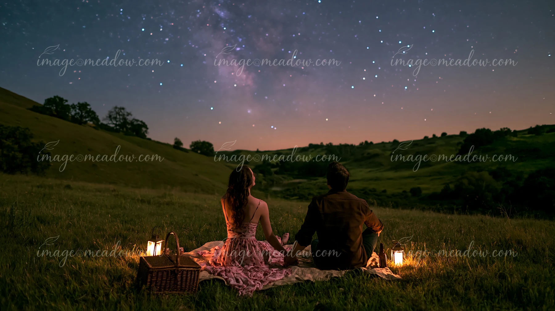 A couple holding hands beneath the night sky sits on grass, surrounded by a soft glow from lanterns.