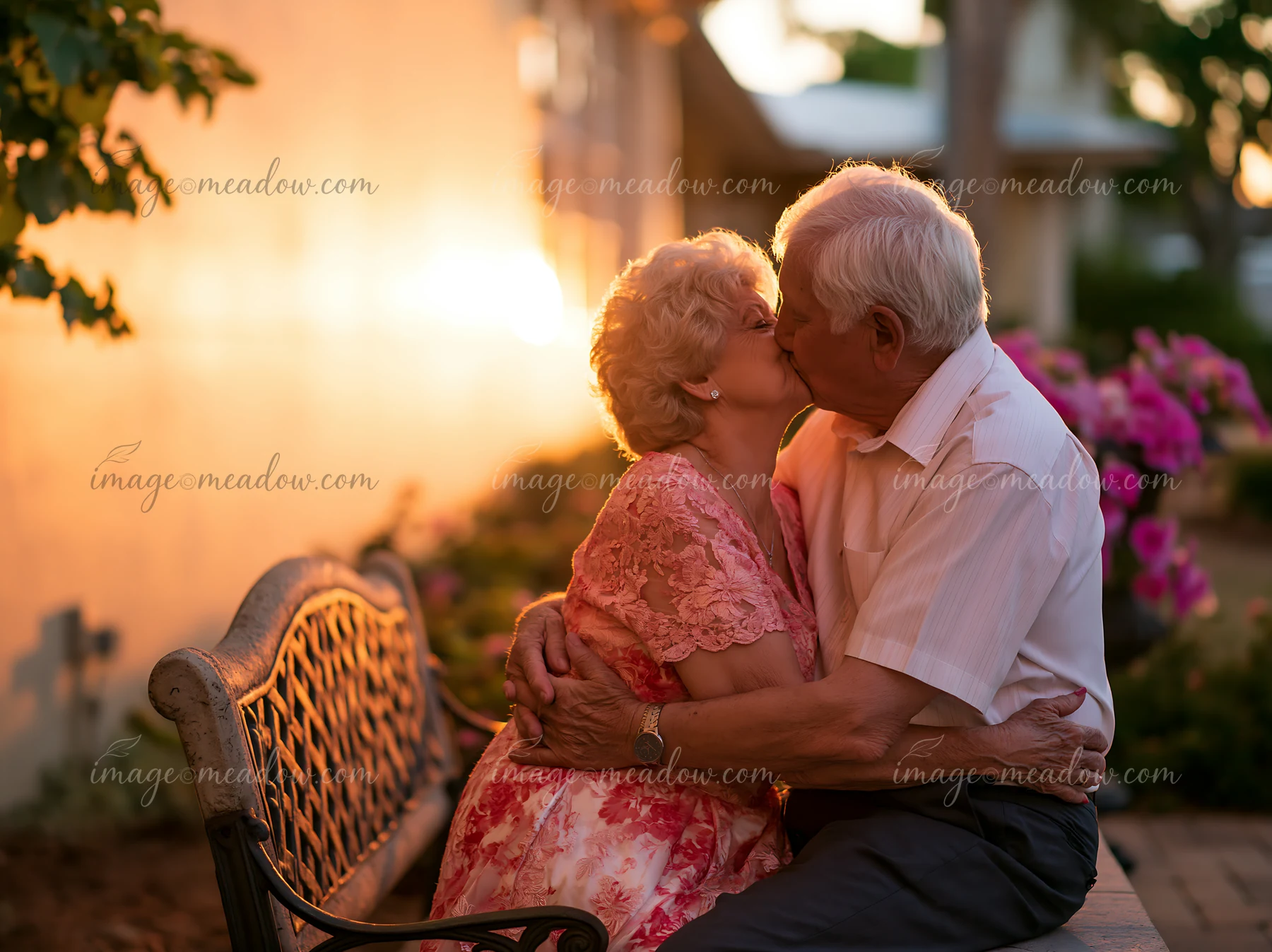 A couple kisses on a bench as the sun sets, surrounded by flowers.