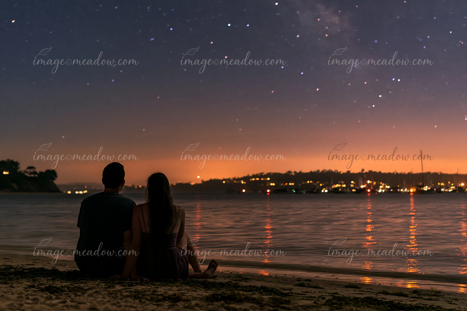 A couple sits together on the beach under stars, watching the night sky.