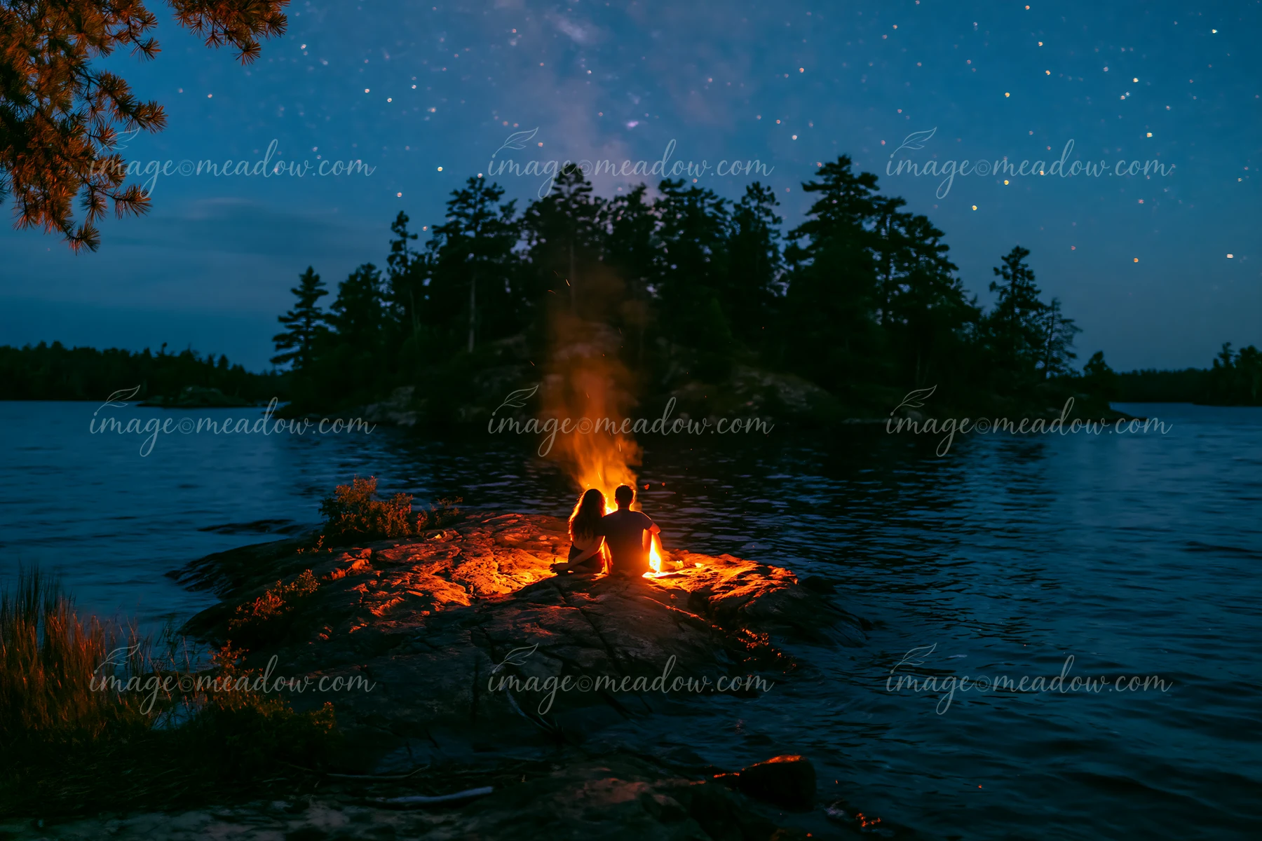 A couple sitting beside a glowing campfire under stars shares a quiet moment on a rocky lakeshore.