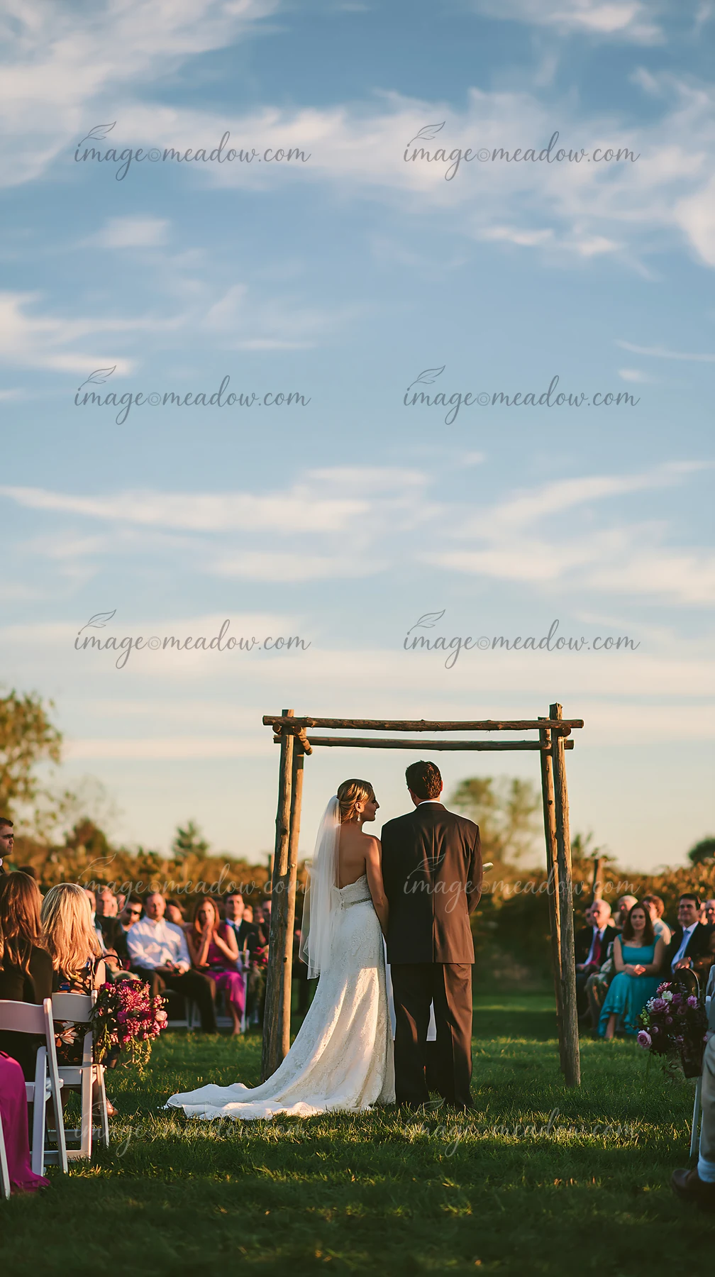 A couple stands closely under a wedding arch during an outdoor ceremony, surrounded by guests seated on chairs.